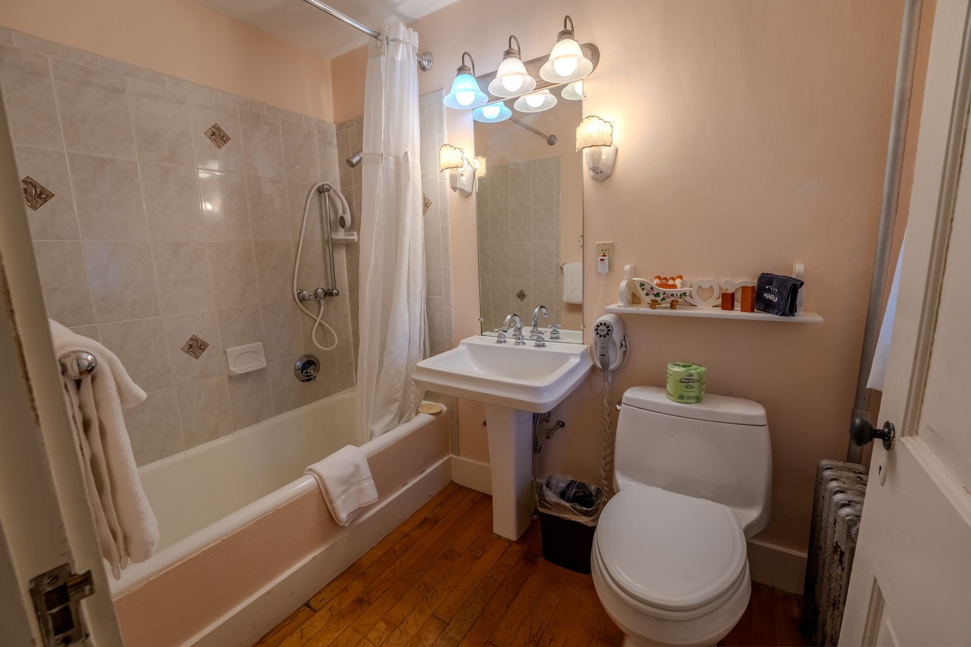 Bathroom with a bathtub, sink, and toilet. Pink walls, wood floor, and a lit mirror.