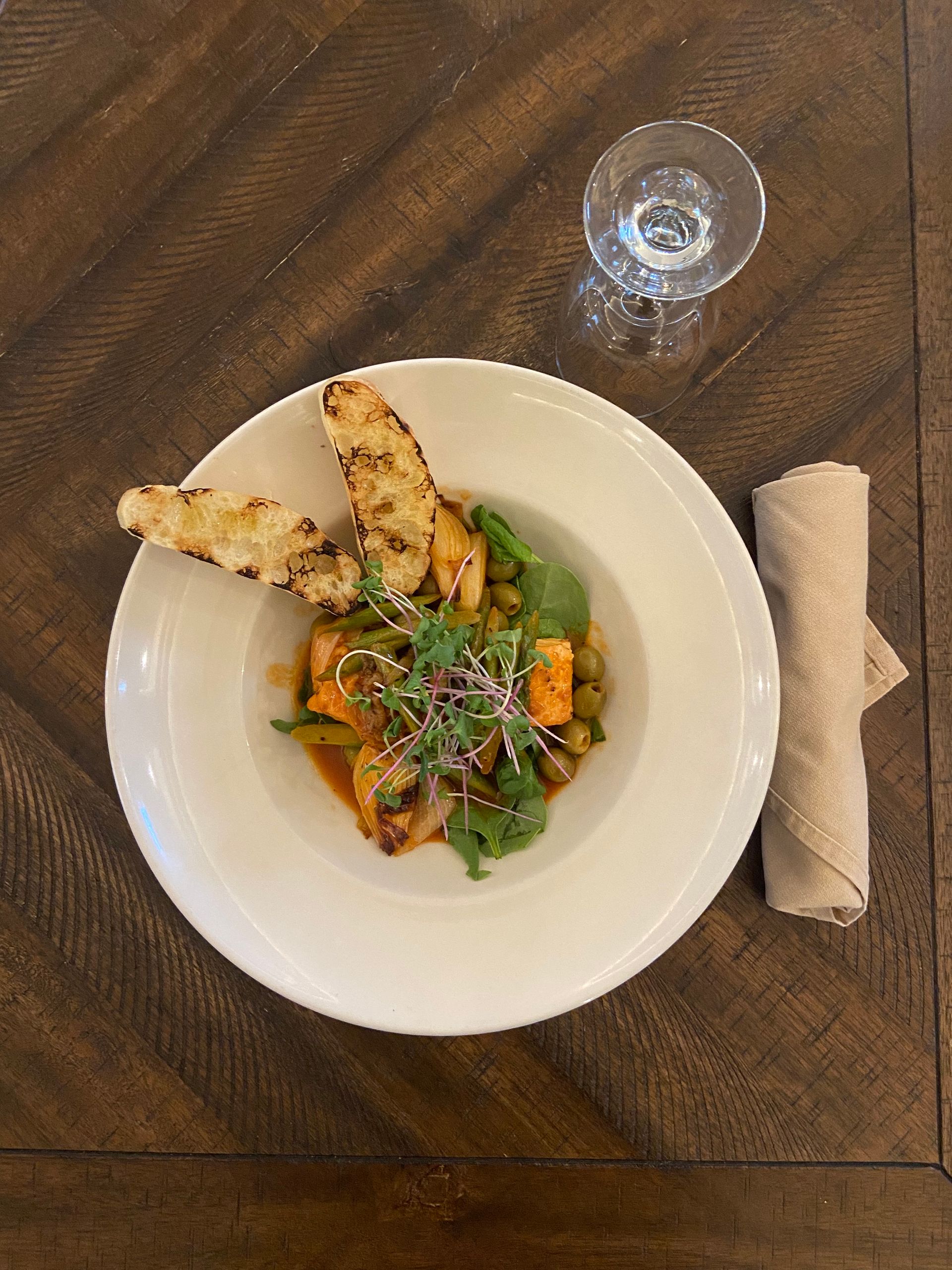 White bowl with a stew and grilled bread, next to a glass and rolled napkin on a wooden table.