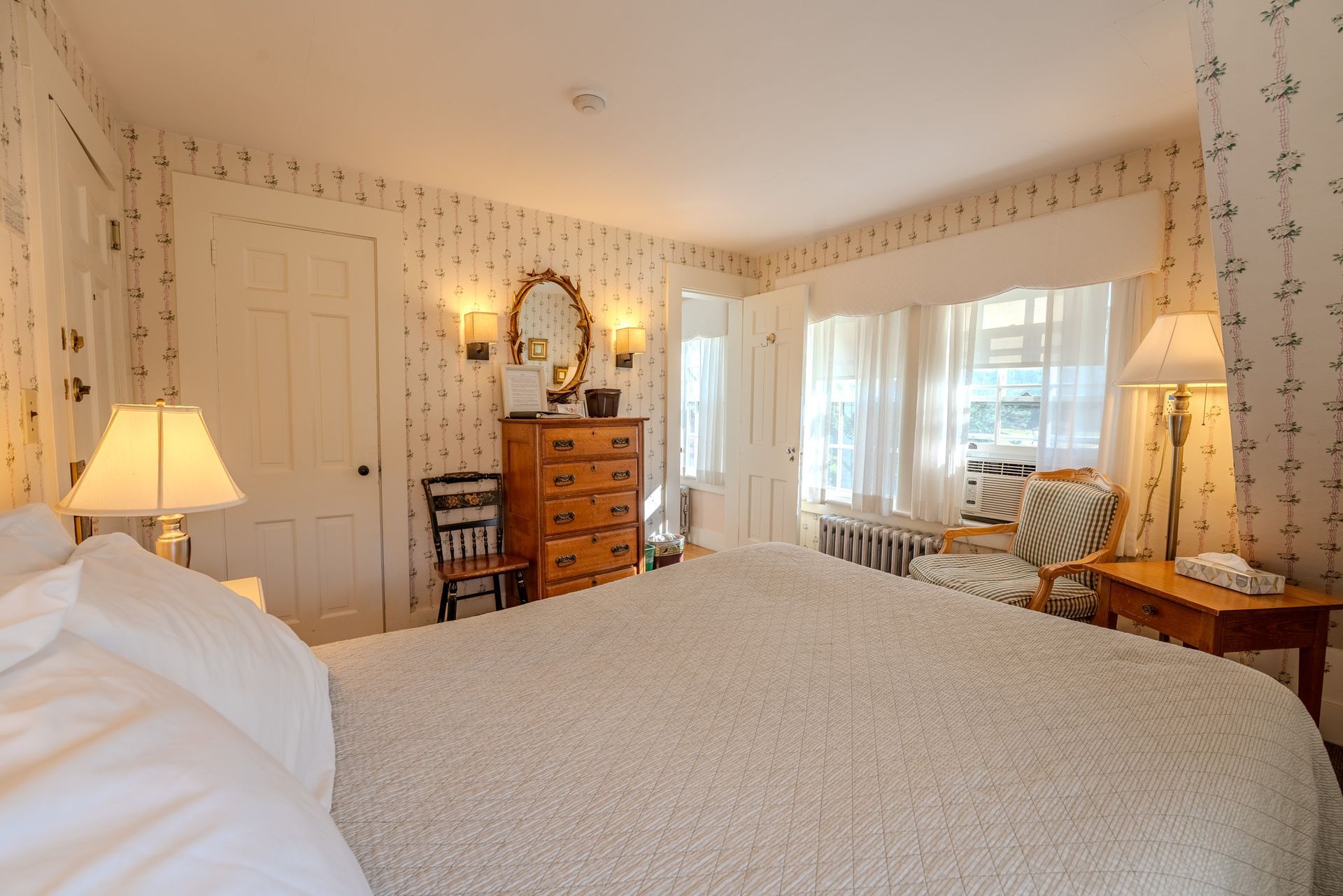 Cozy bedroom with floral wallpaper, a large bed, a dresser, and a window with a sitting area.