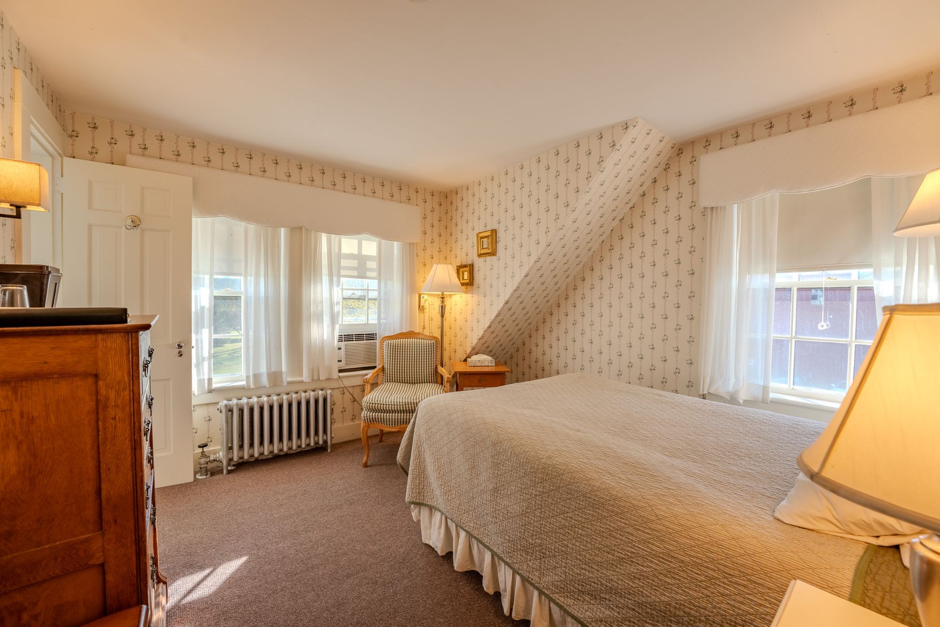 Bedroom with bed, windows, patterned wallpaper, antique dresser and lamps.