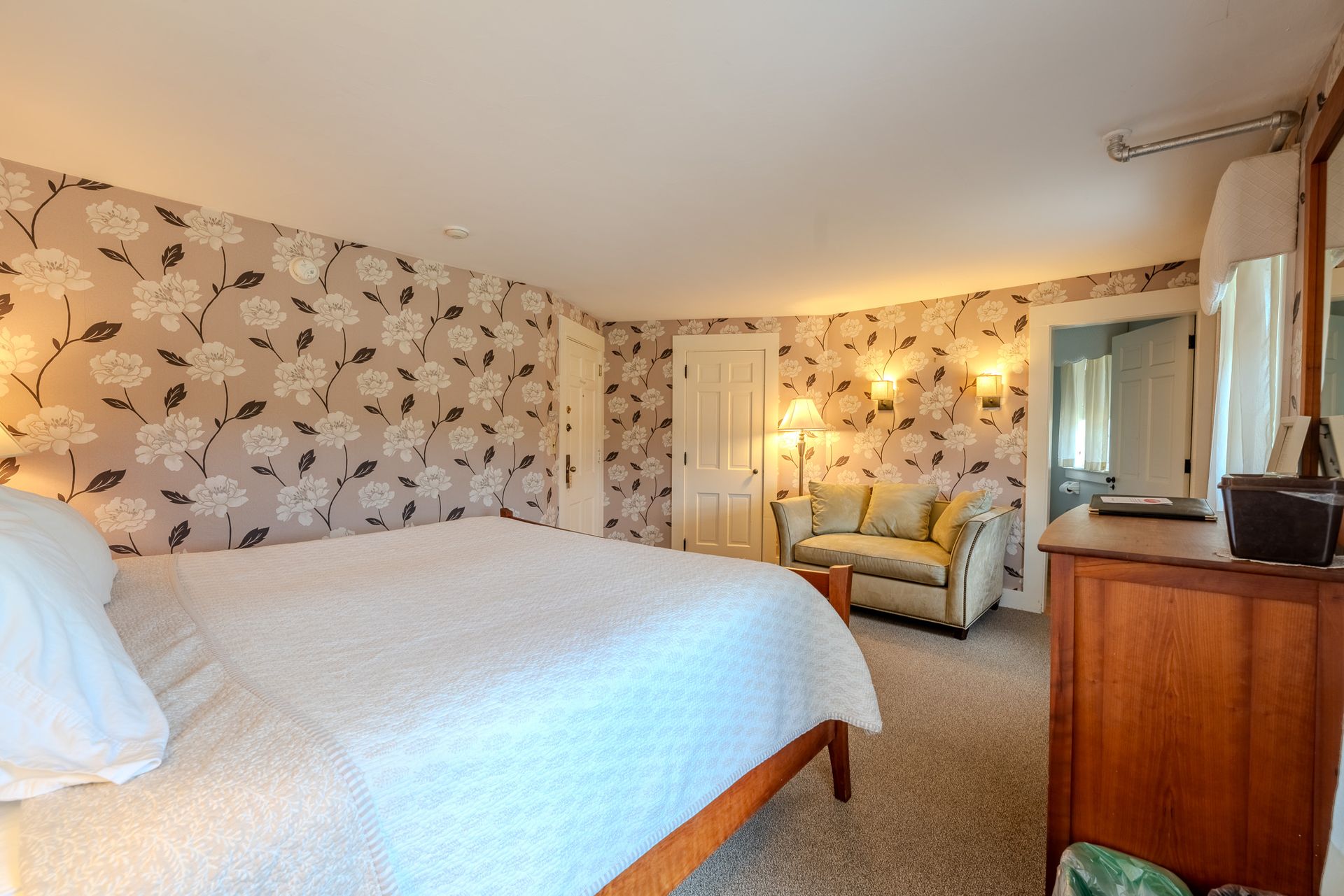 Bedroom with floral wallpaper, bed, armchair, dresser, and door.