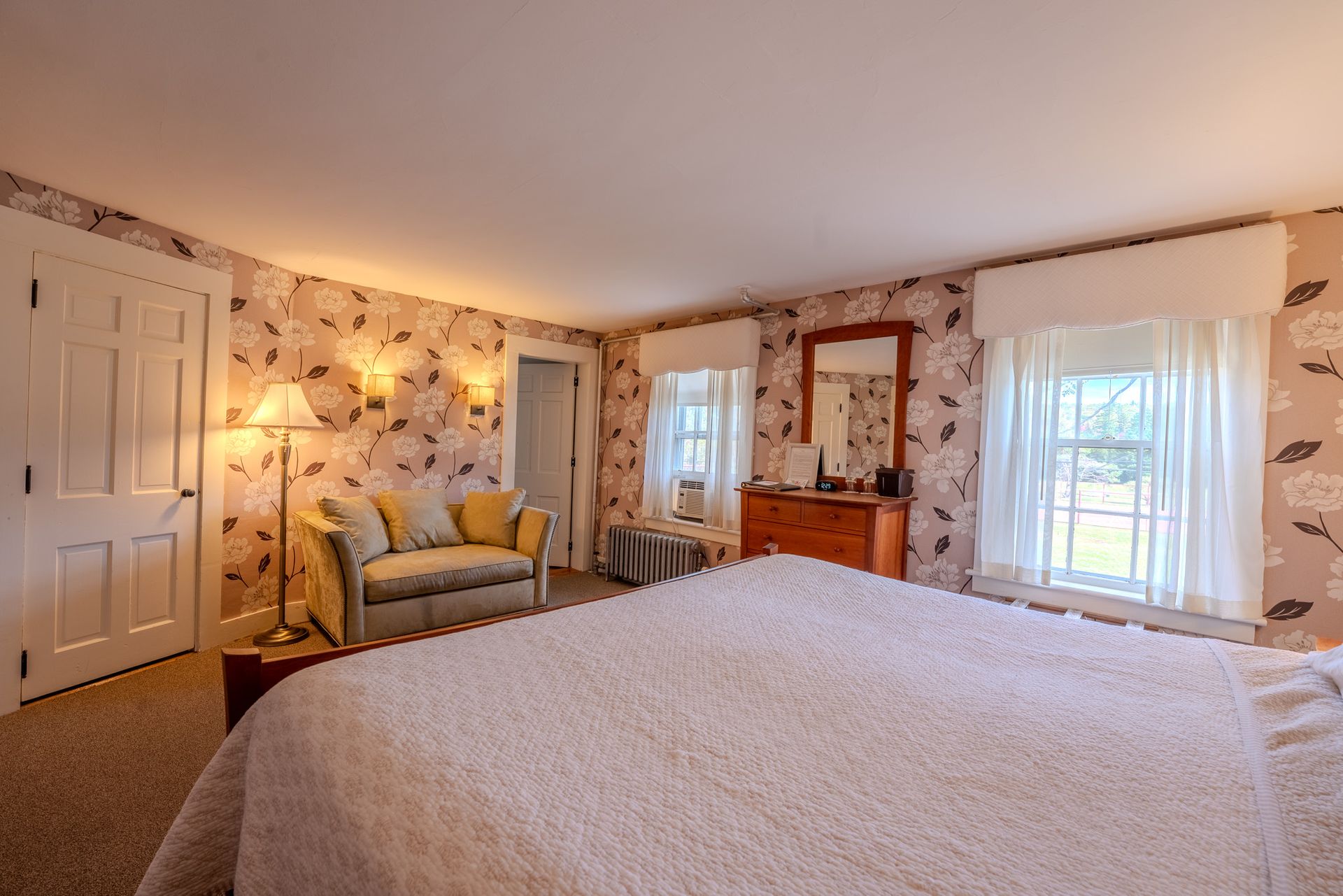Bedroom with floral wallpaper, queen bed, loveseat, dresser, windows, and a closed door.