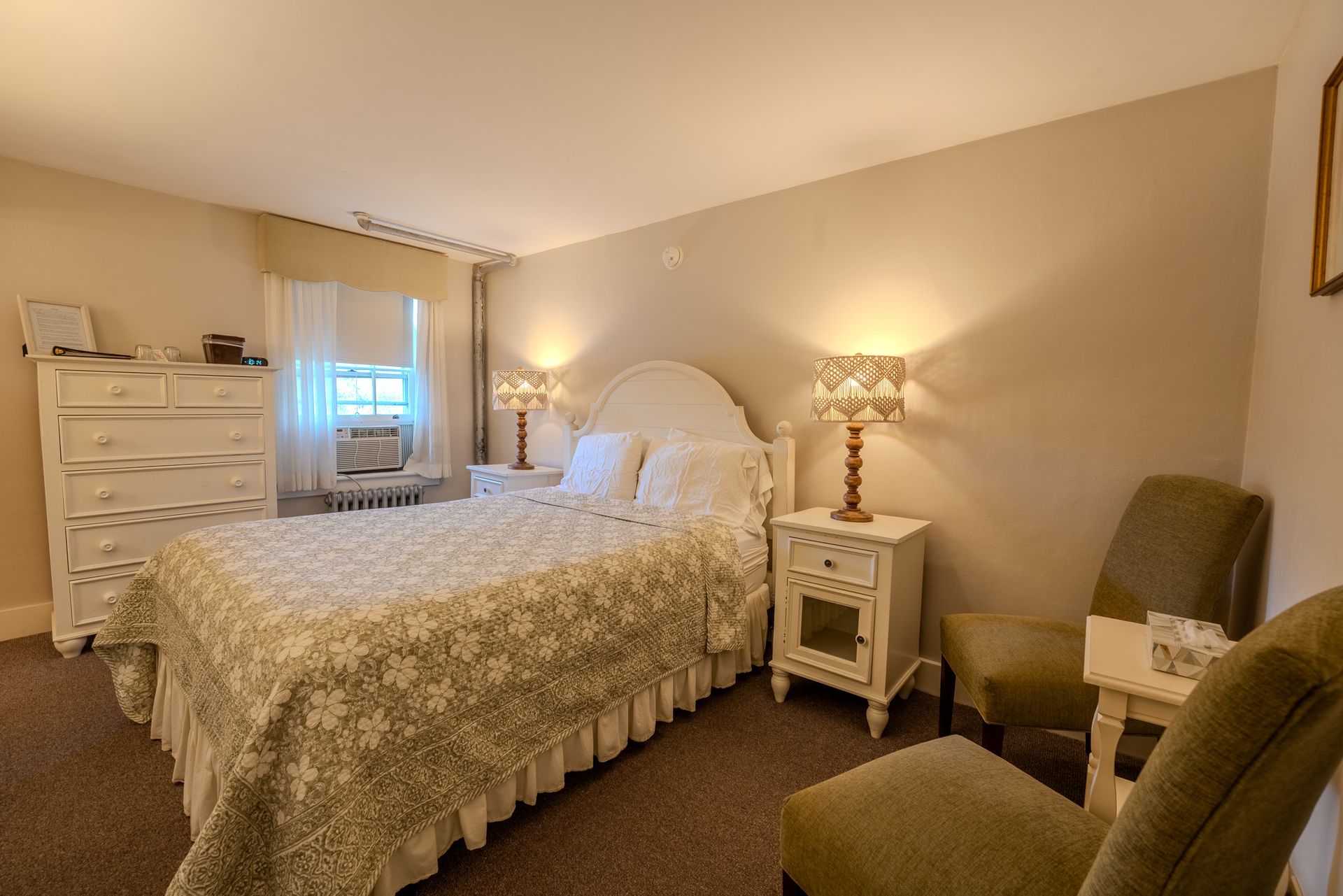 Bedroom with a bed, two chairs, two nightstands, and a dresser. Beige walls and carpet.