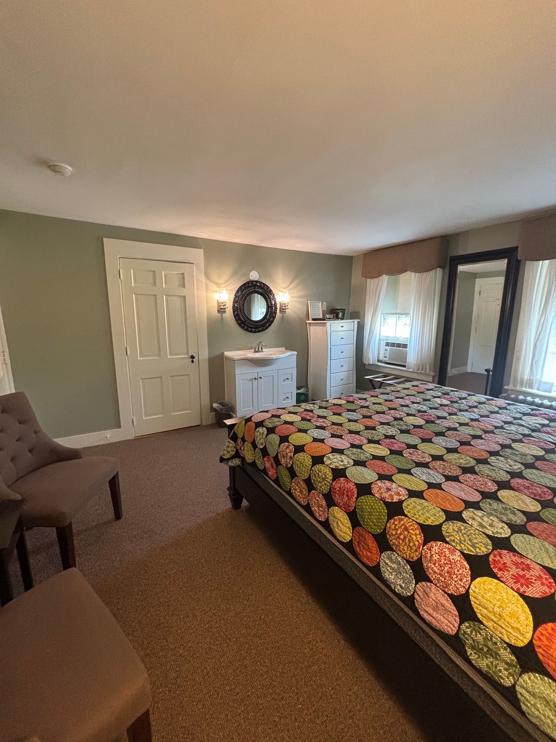 Bedroom with bed covered in colorful quilt, white dresser, mirror, and armchairs on brown carpet.