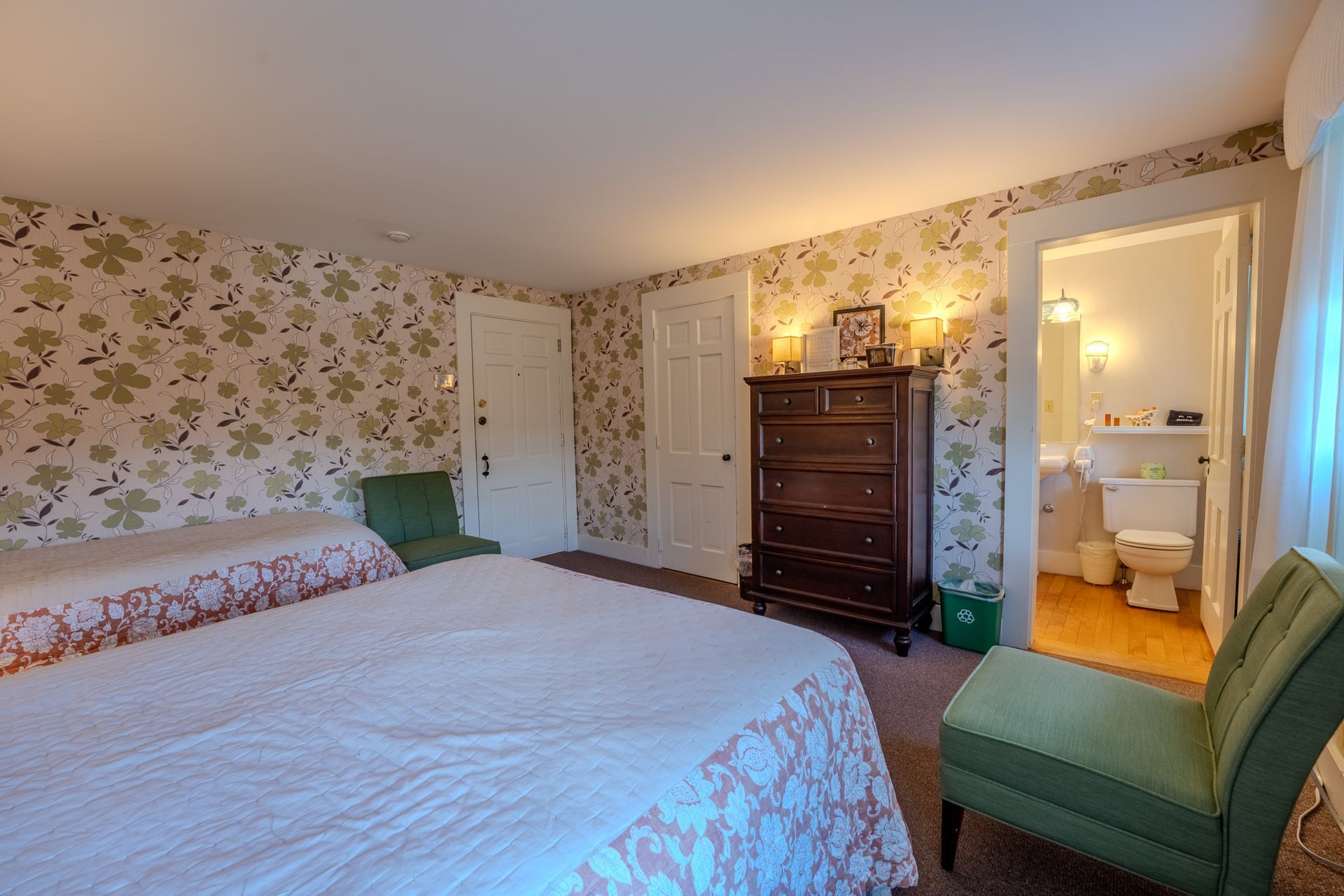 Bedroom with floral wallpaper, two beds, dresser, green chairs, and a bathroom doorway.