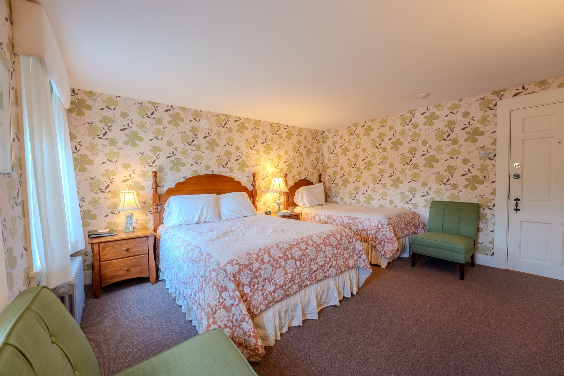 A hotel room with two beds, floral wallpaper, a green chair, and a nightstand with a lamp.