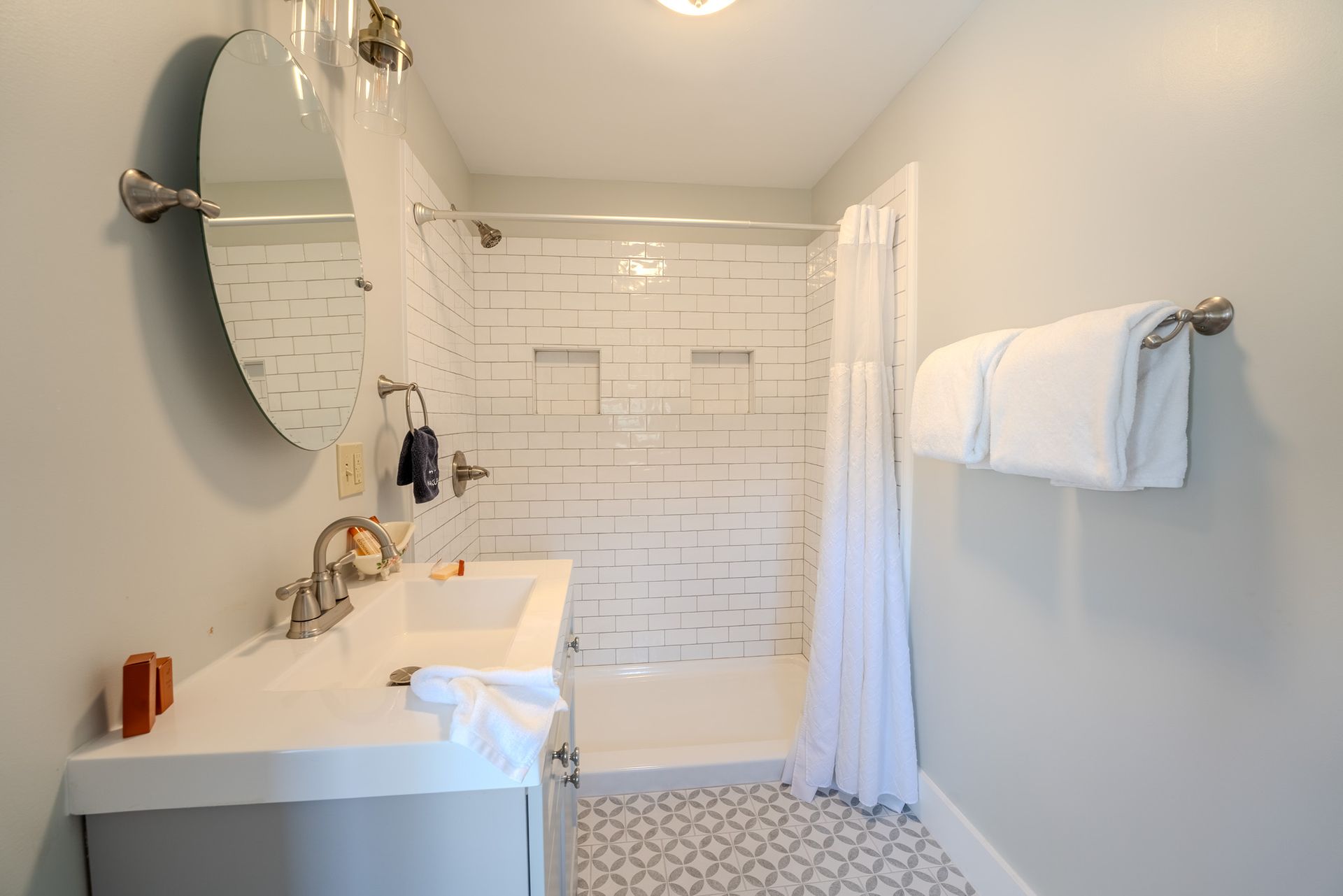 Bathroom with white shower, vanity, and towels. Round mirror, patterned floor.