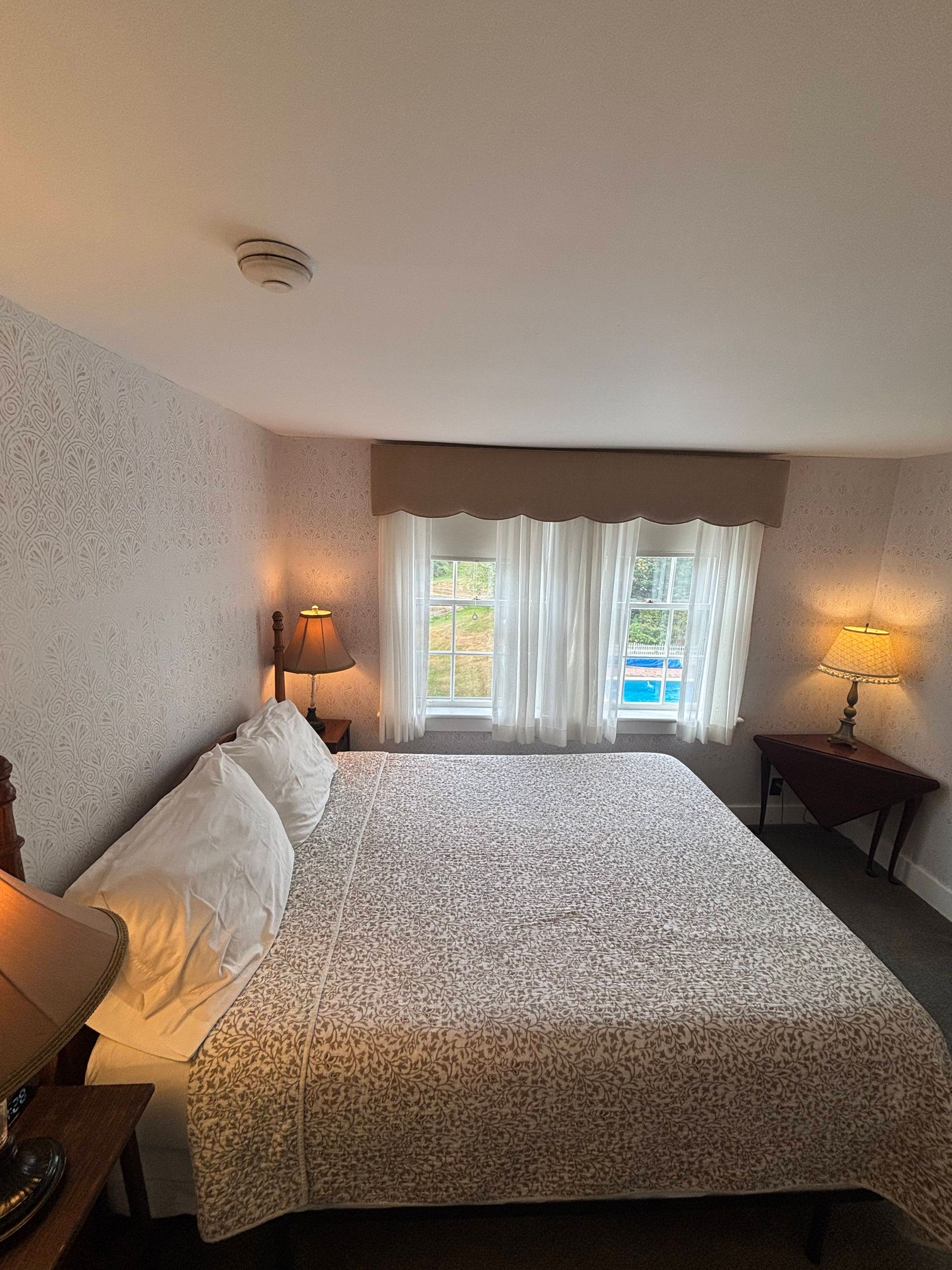 Bedroom with a large bed, two windows with curtains, bedside lamps, and patterned wallpaper.