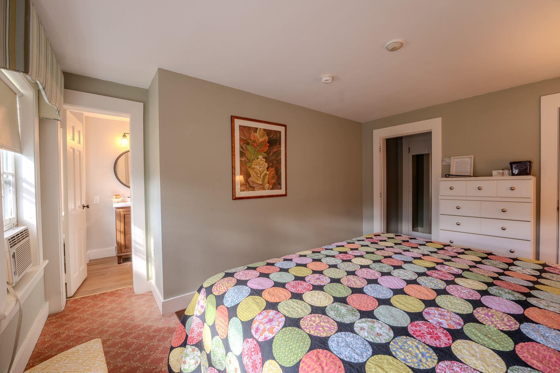 Bedroom with colorful quilt, artwork, doorway to bathroom, white dresser.