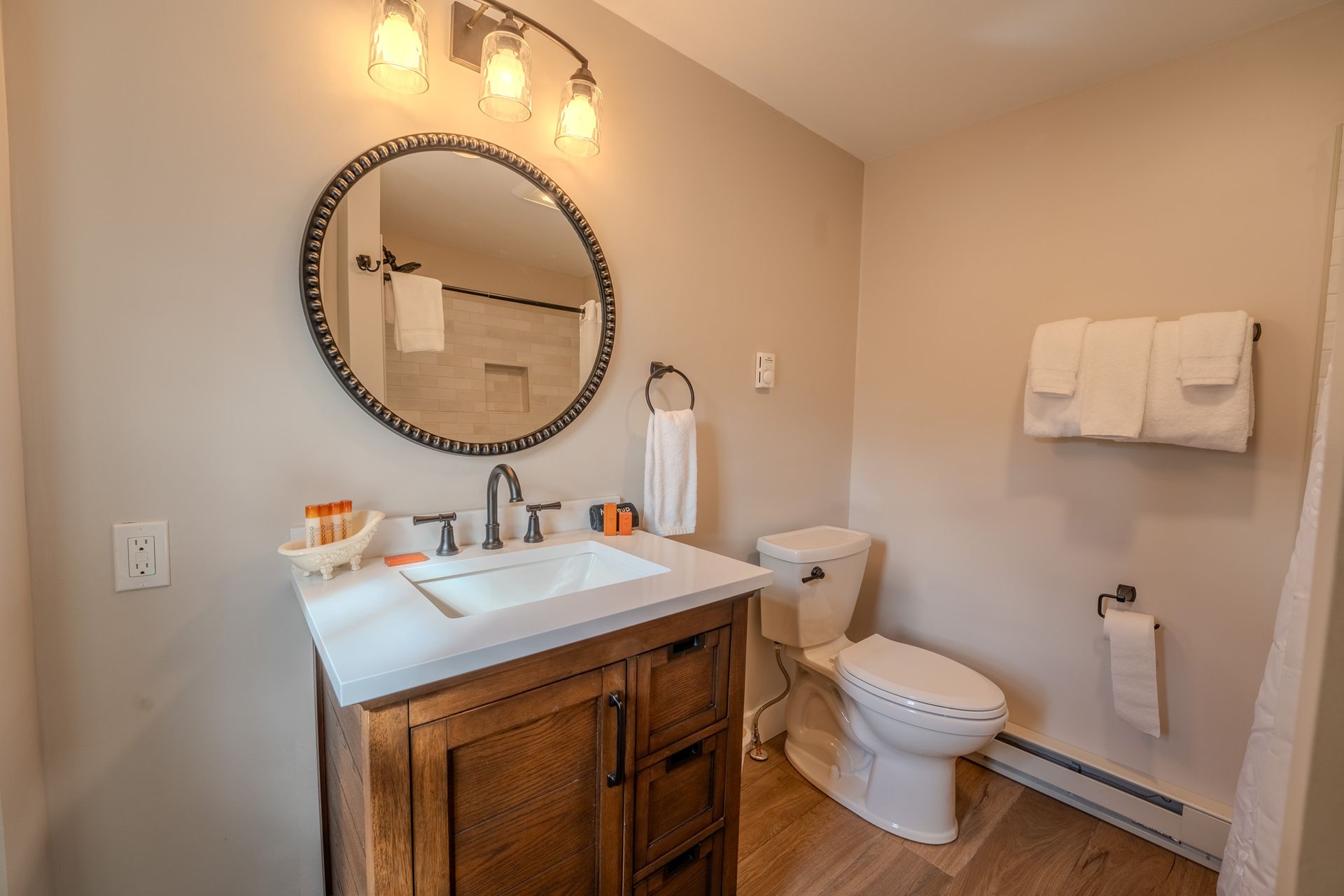 Bathroom with wooden vanity, round mirror, toilet, and towels. Beige walls, wooden floor, and natural light.