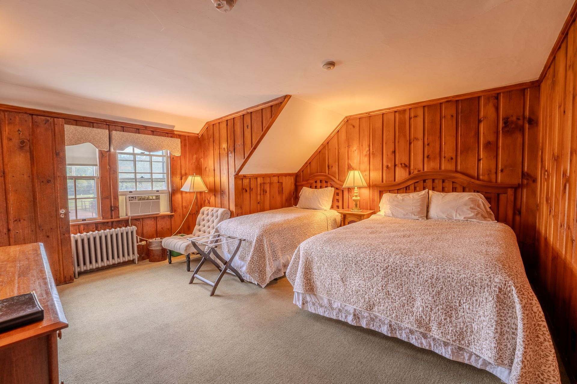 A rustic hotel room with wood paneling, two beds, and a chair near windows.
