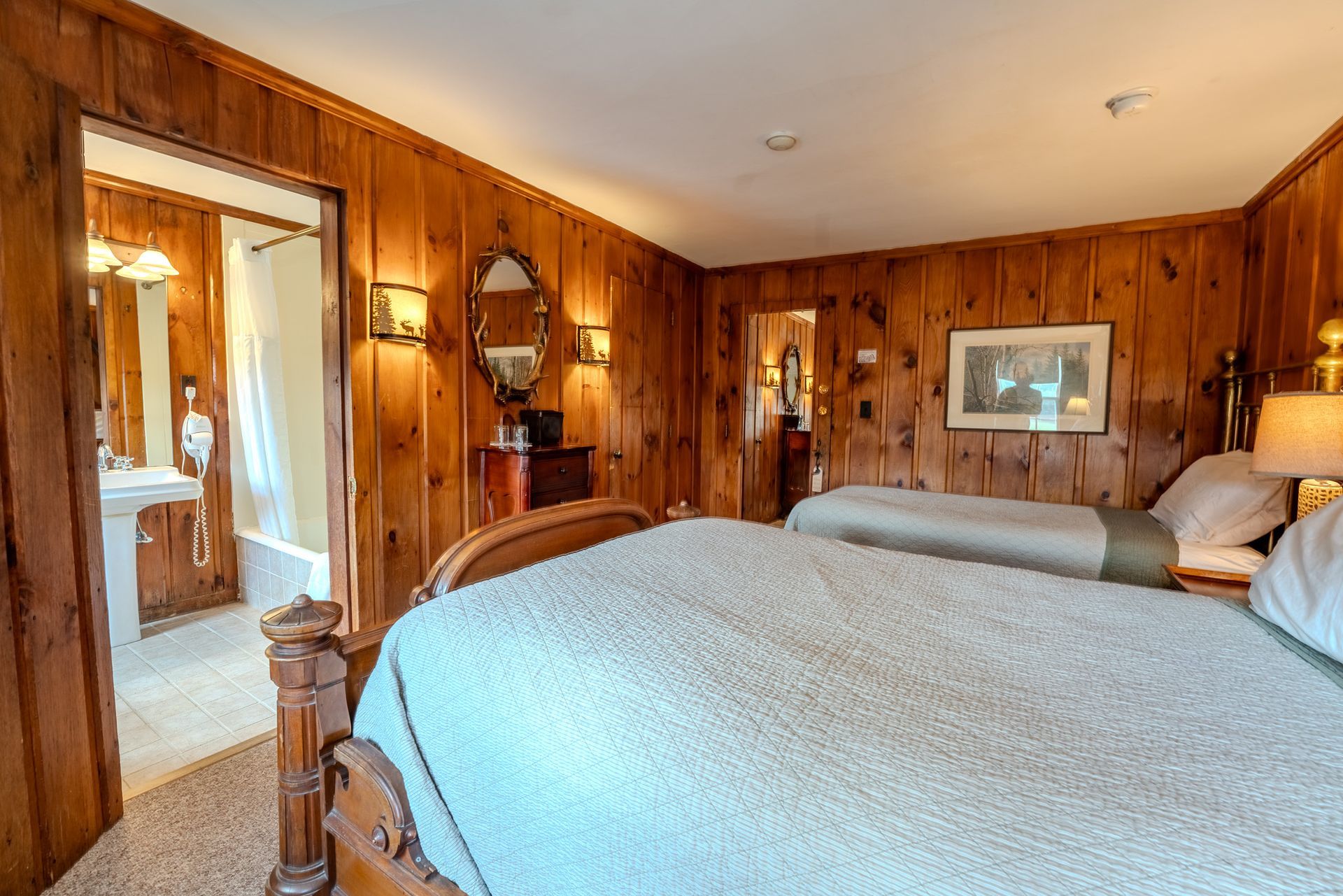 A bedroom with wood paneling, two beds, and a bathroom visible in the doorway.