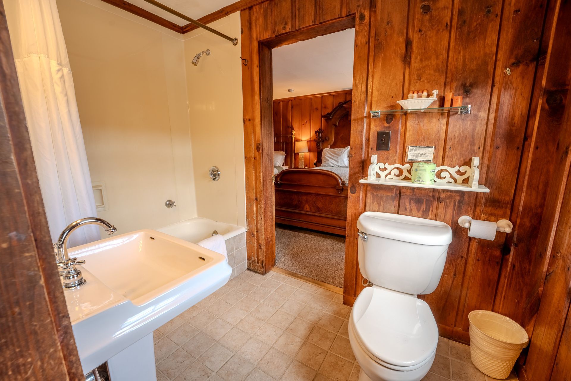 Bathroom with wood paneling, a toilet, sink, and doorway to a bedroom with a bed.