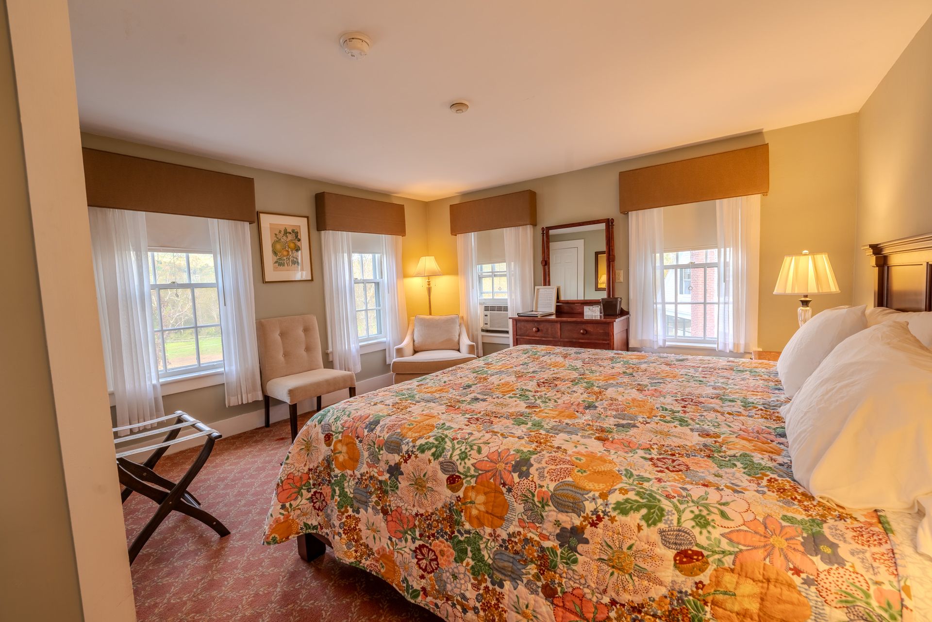 Bedroom with a large bed, windows, chairs, and a dresser. Light yellow walls, floral quilt, and soft lighting.