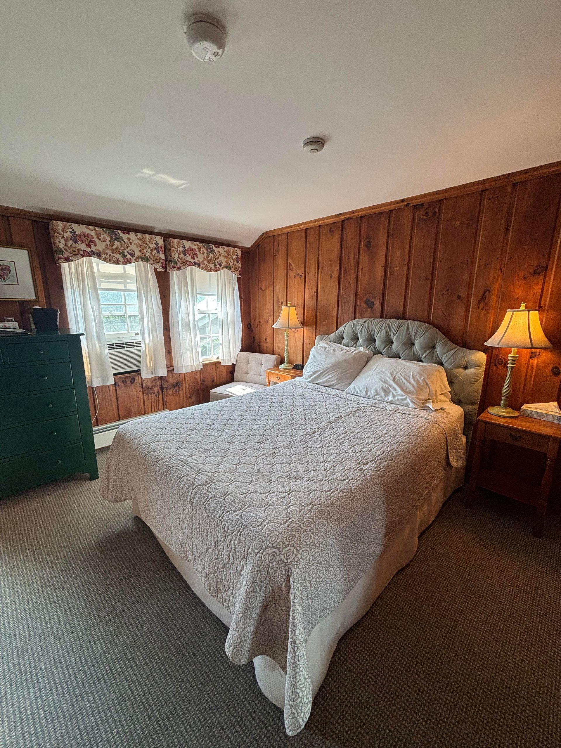 Bedroom with wood-paneled wall, a bed with a floral patterned quilt, and small windows with curtains.
