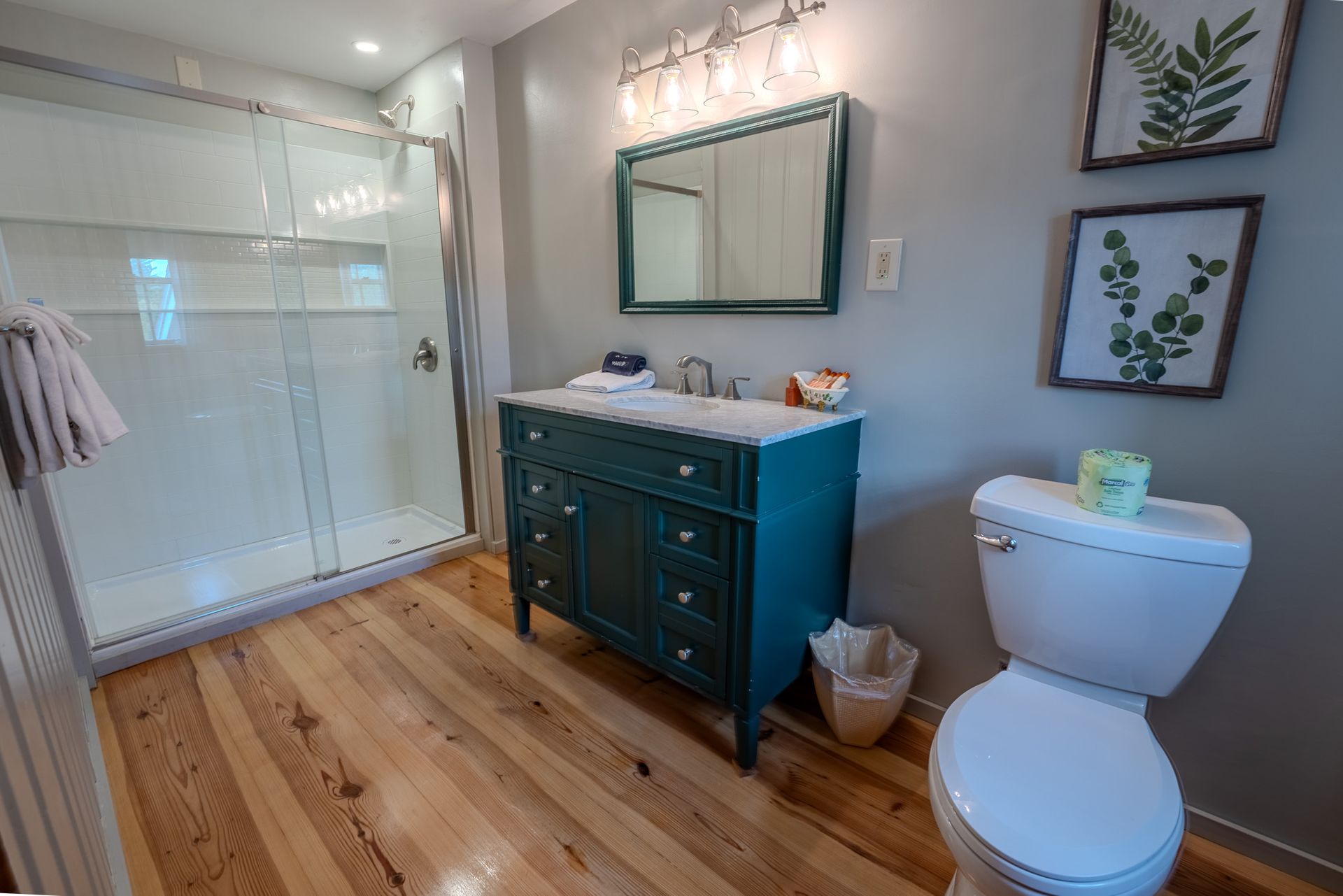 Bathroom with teal vanity, glass shower, wooden floor, and botanical art.