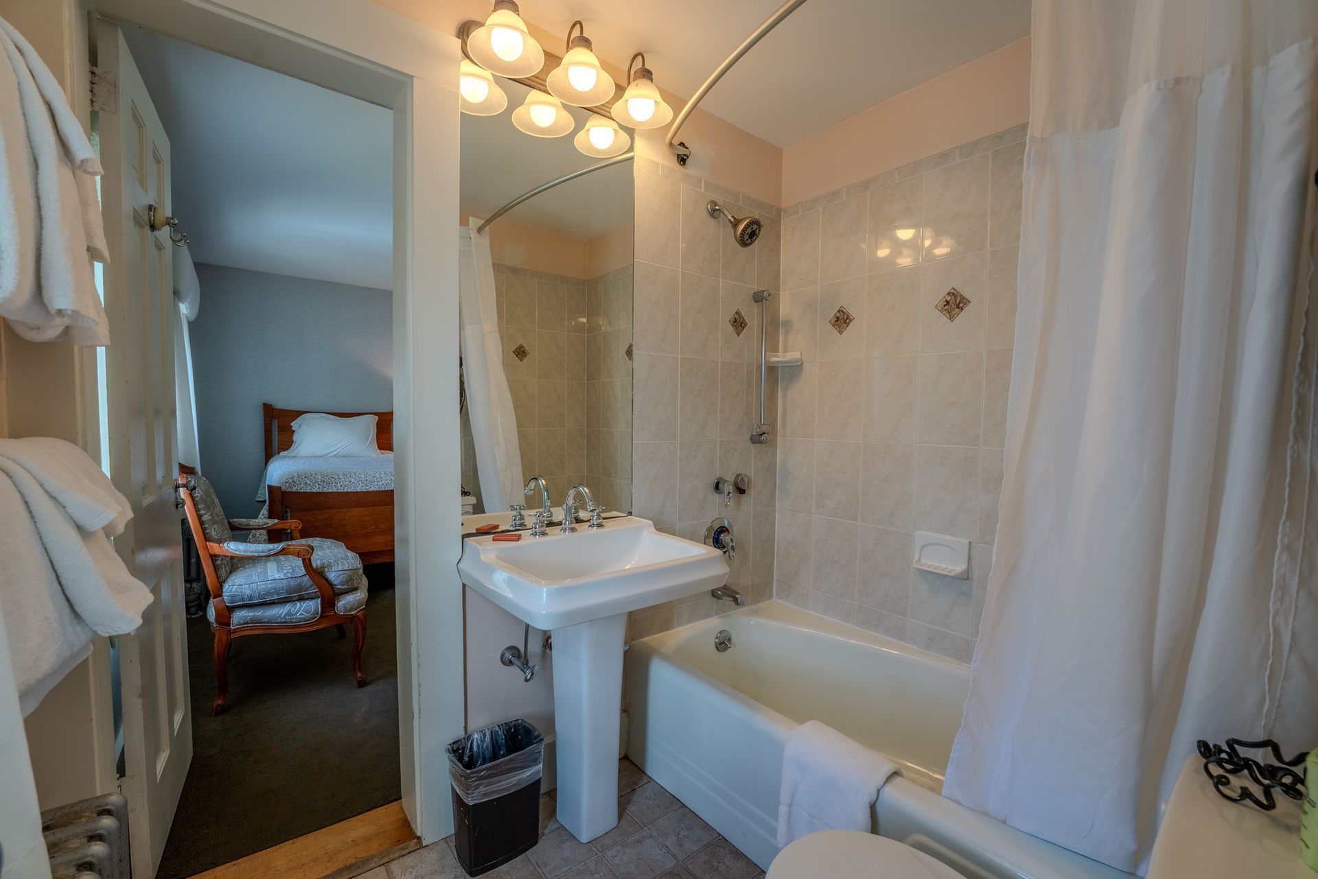 Bathroom with a white pedestal sink, bathtub, and view into a bedroom with a bed and chair.