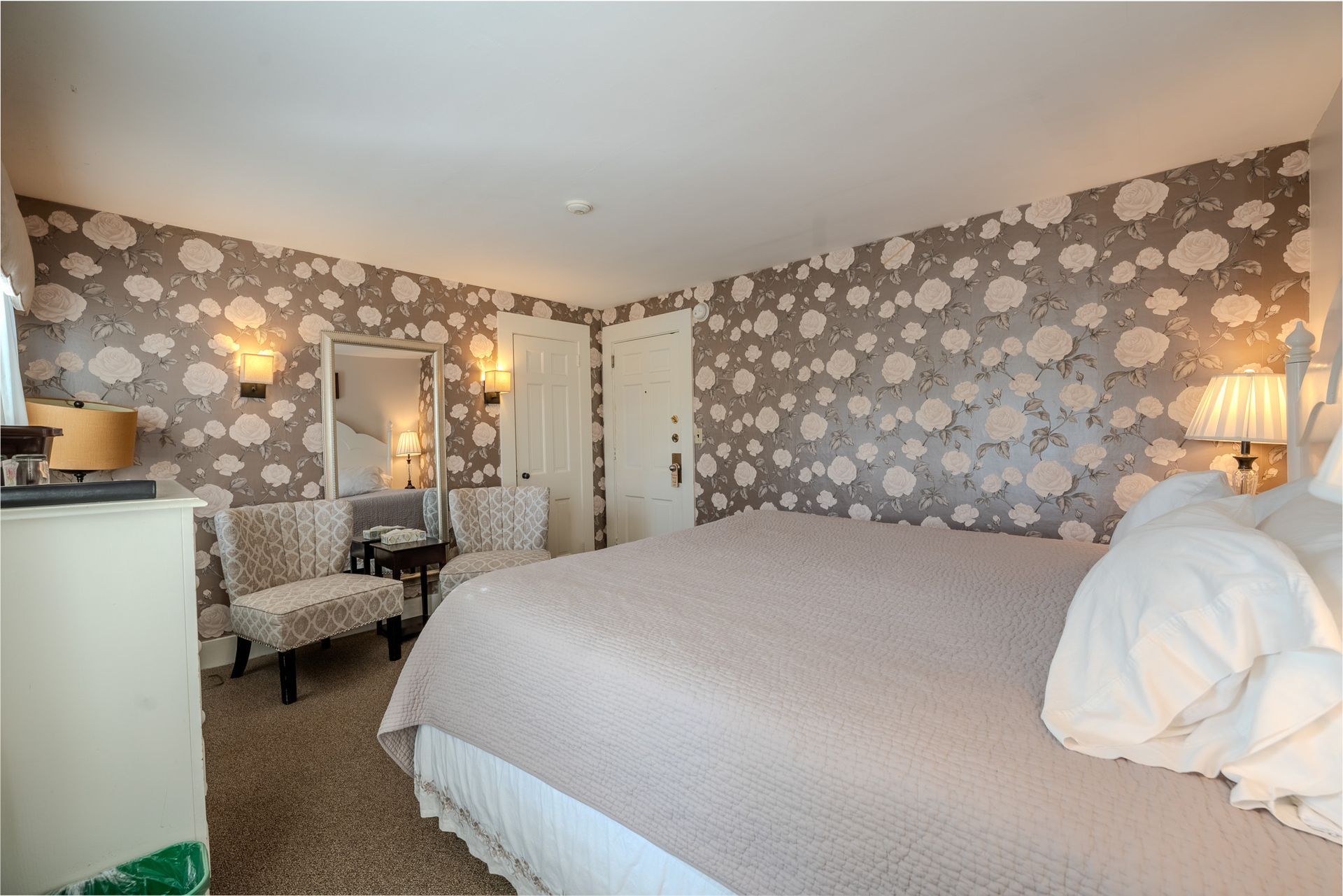 Bedroom with floral wallpaper, a bed, two chairs, a mirror, and a white dresser.