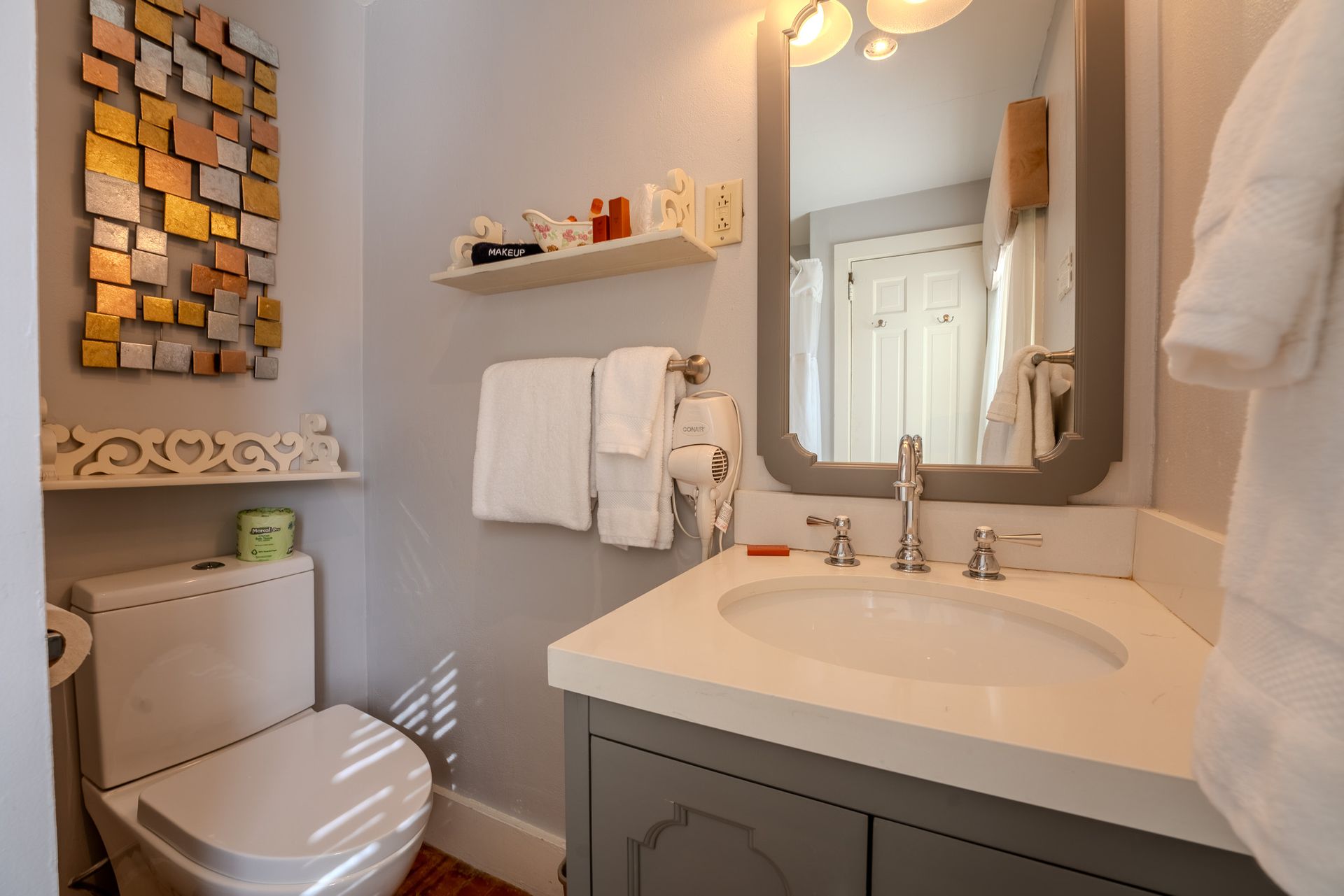 Bathroom with gray vanity, white sink, toilet, mirror, and towel rack; an art piece hangs on the wall.