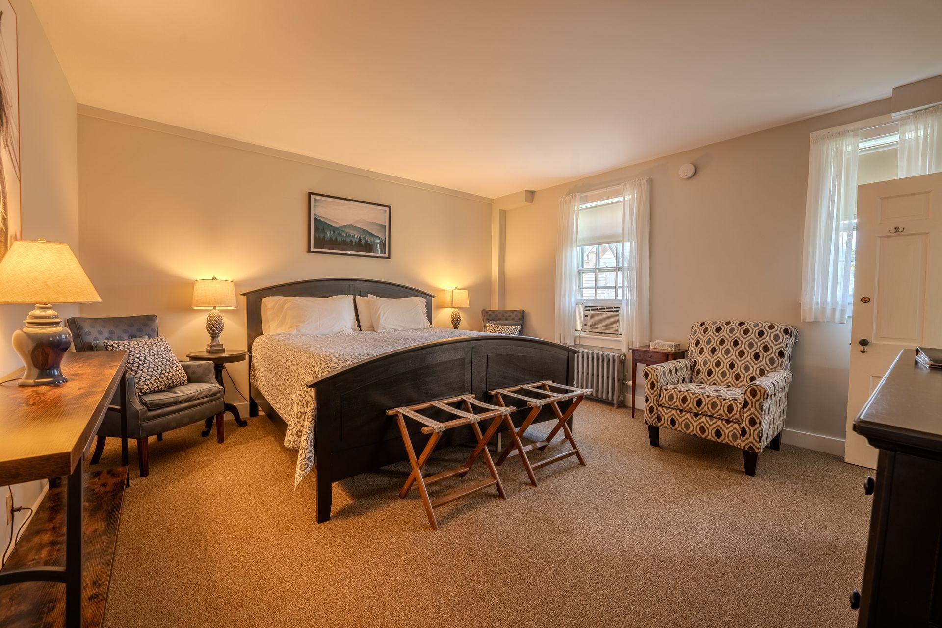 Bedroom with a dark bed frame, luggage racks, armchairs, lamps, and a desk.