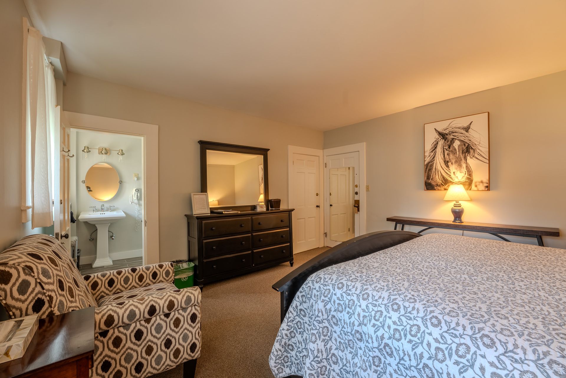 Bedroom with bed, dresser, armchair, artwork, and an ensuite bathroom.