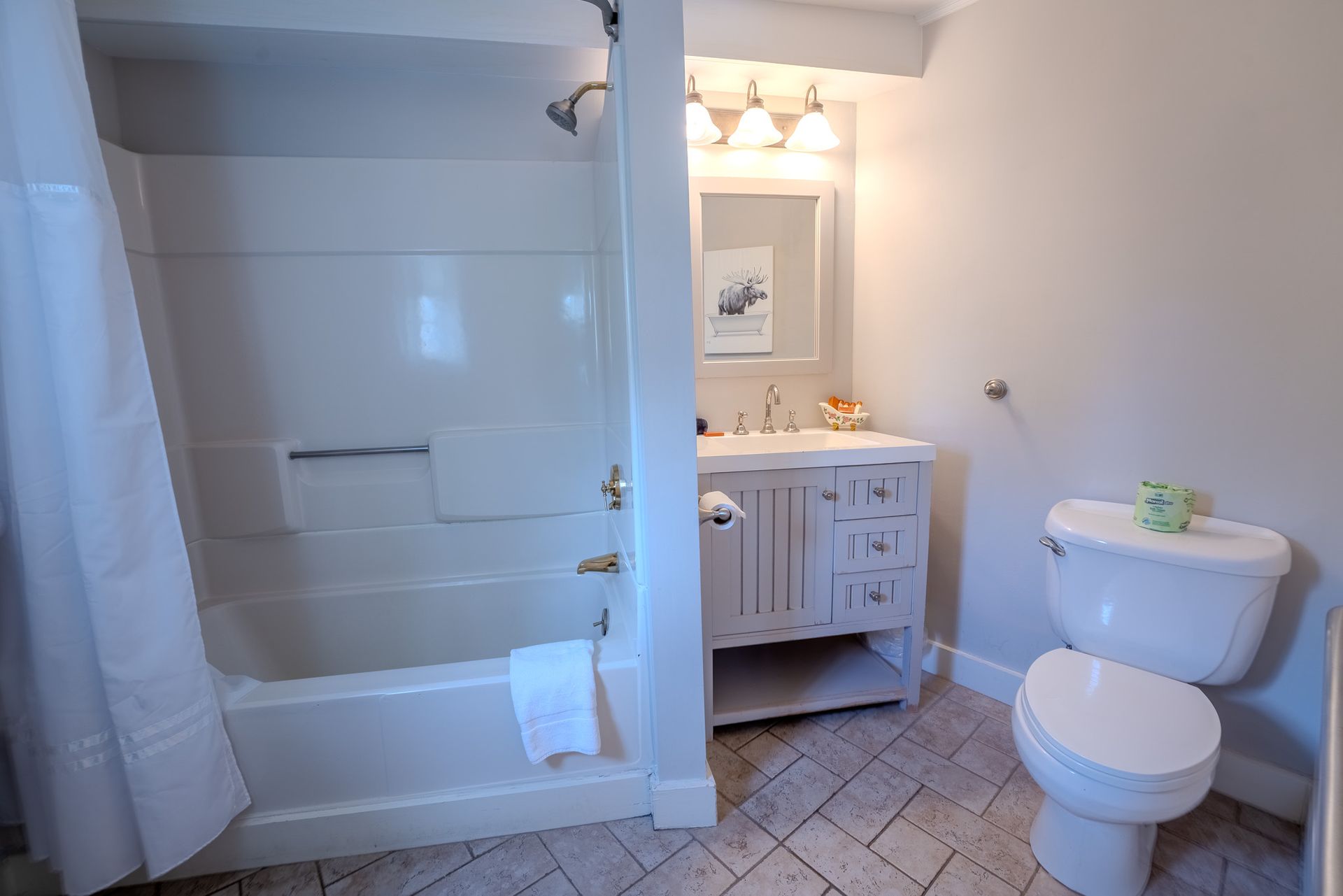 A small, bright bathroom with a tub/shower, vanity, and toilet. Beige walls, tile floor, and white fixtures.