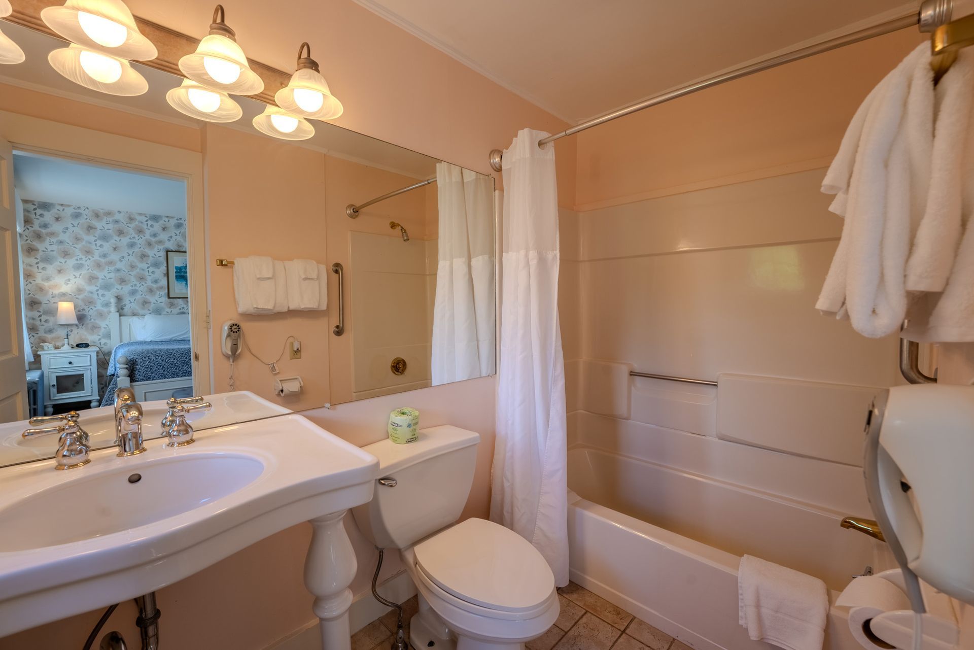 Bathroom with sink, toilet, and bathtub.  Pink walls, white towels, and mirror reflecting into a bedroom.