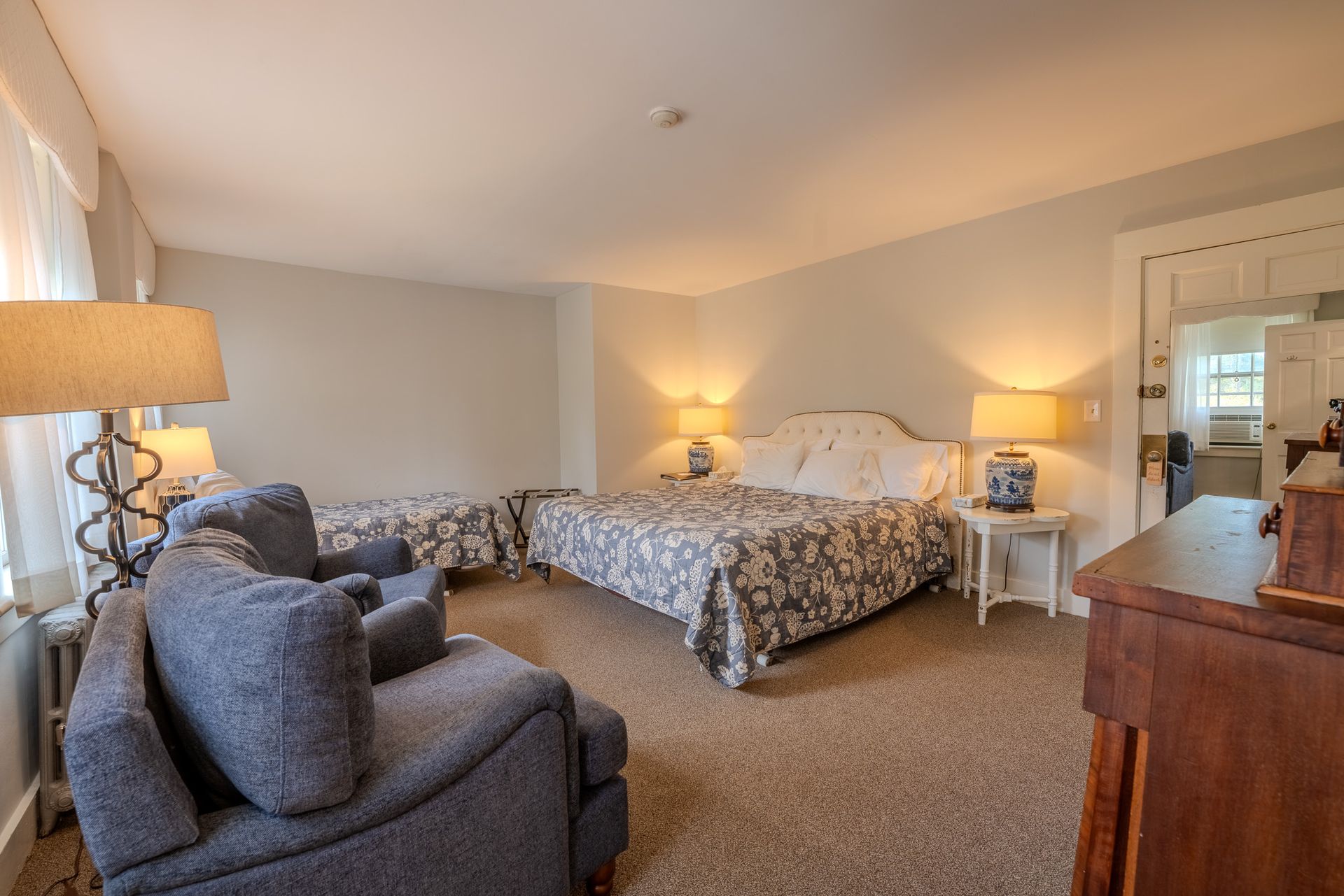 Bedroom with bed, loveseat, and two chairs, with lamps and doorway to another room, neutral color scheme.