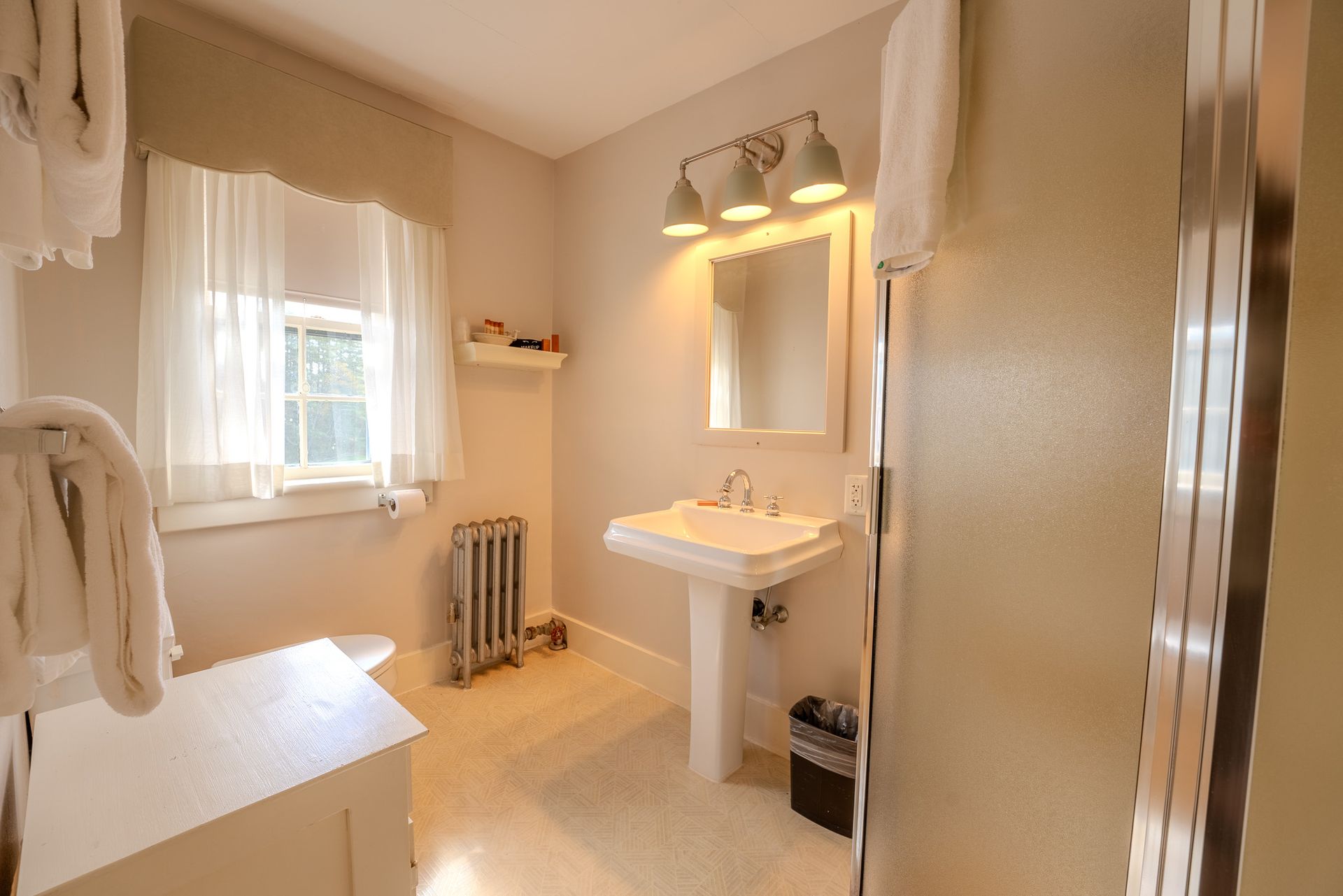 Bathroom with pedestal sink, shower, window, and radiator. Beige walls and floor.