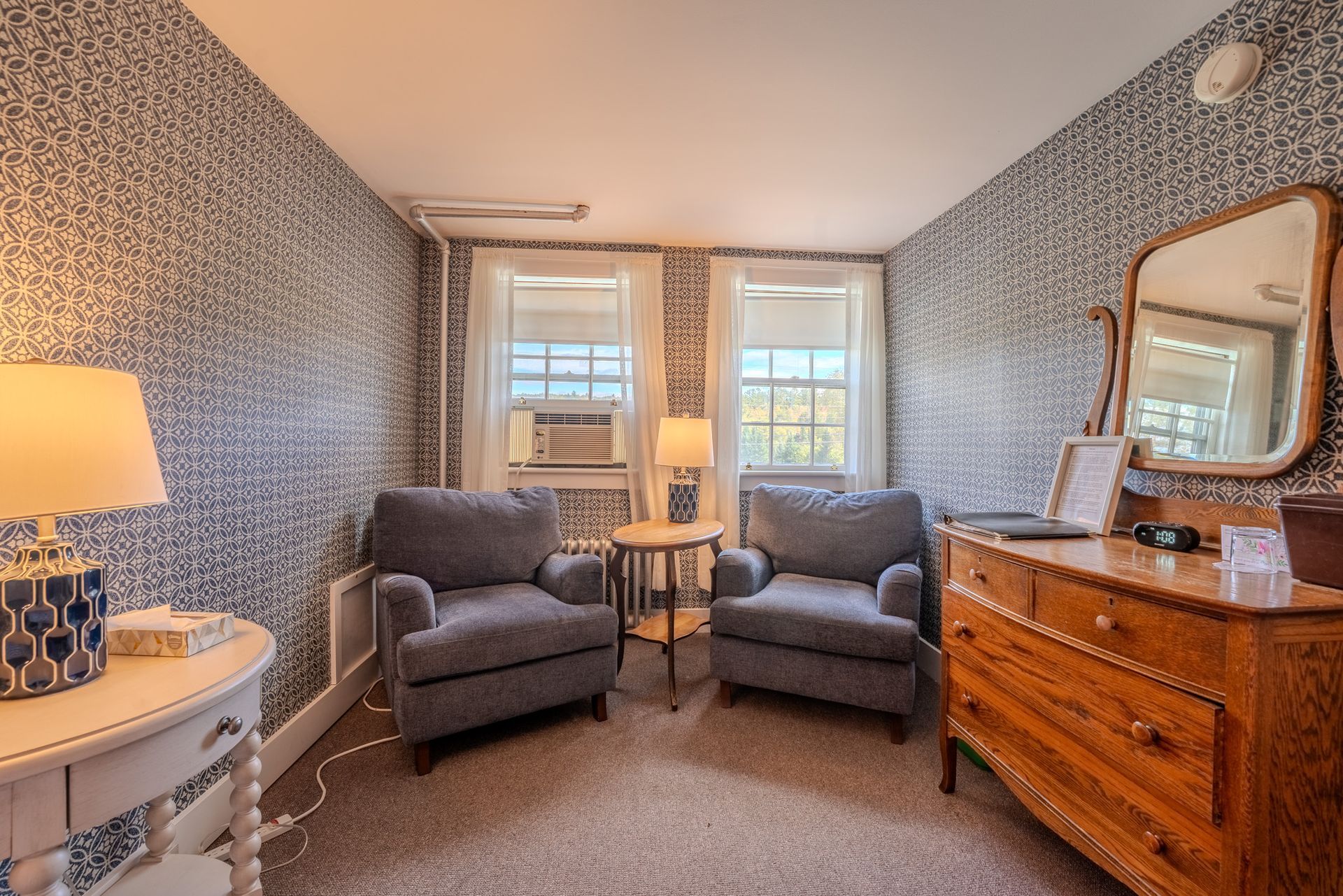 A cozy bedroom with two gray armchairs, antique dresser, and patterned wallpaper.