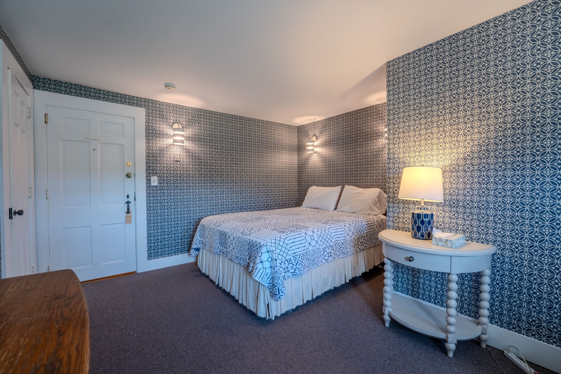 Bedroom with patterned wallpaper, bed, nightstand, and closed door. Blue and white tones.