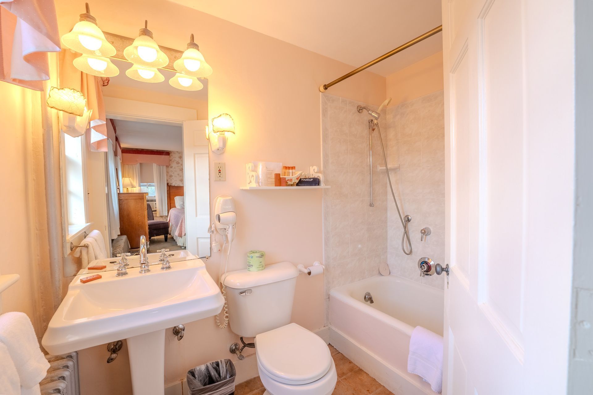 Bathroom with a white pedestal sink, toilet, and bathtub, soft pink walls, and a glimpse into a bedroom.