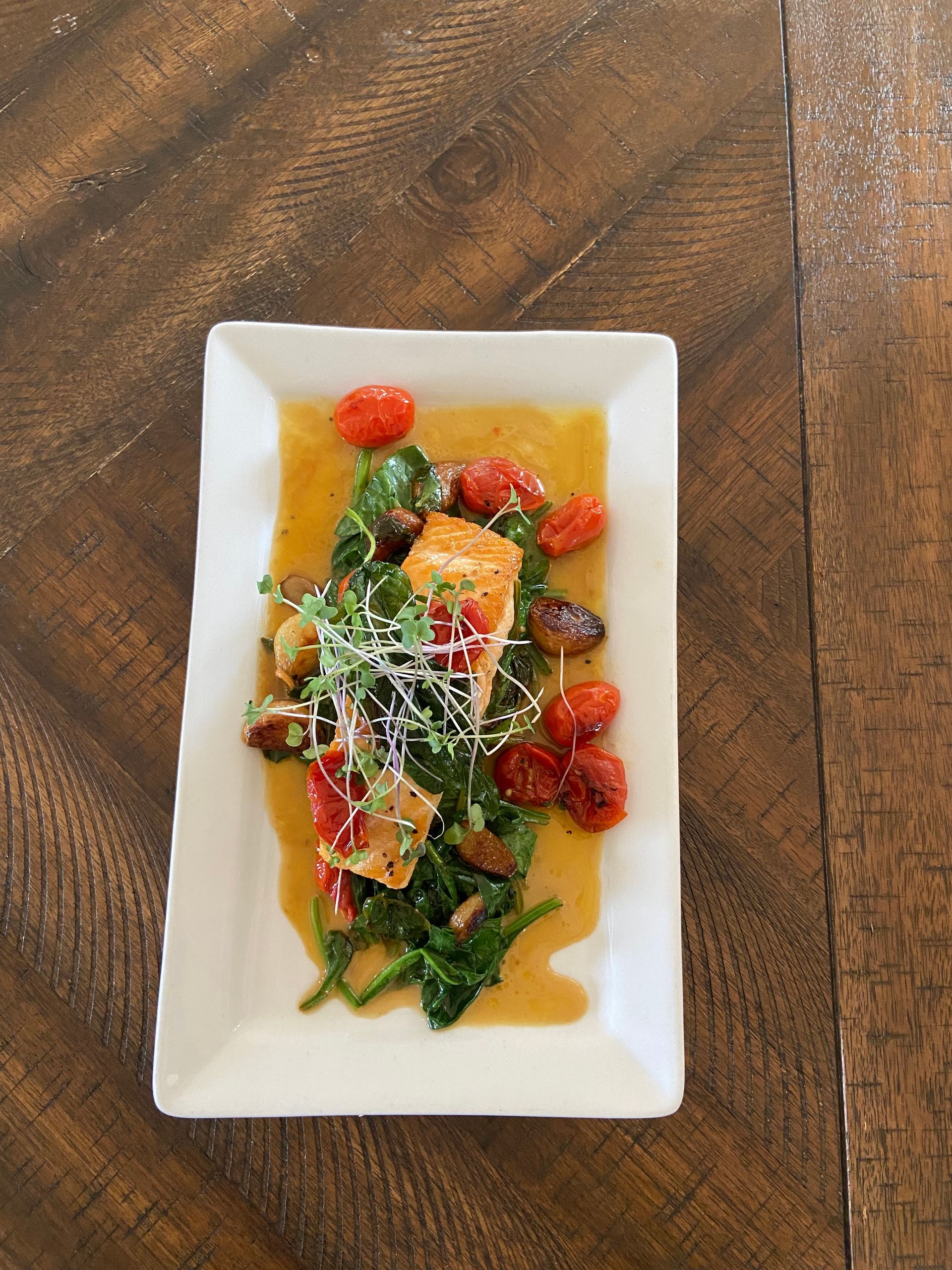 Salmon dish with cherry tomatoes, spinach, and sprouts on a white rectangular plate, on a wooden table.