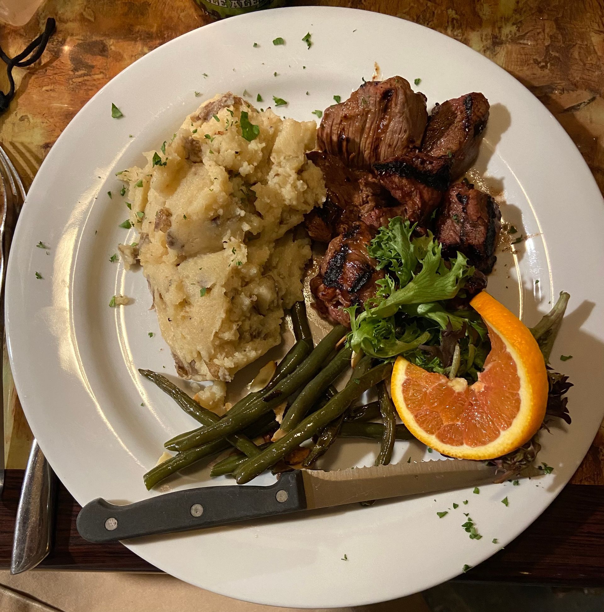Steak bites with mashed potatoes, green beans, and an orange slice on a white plate.