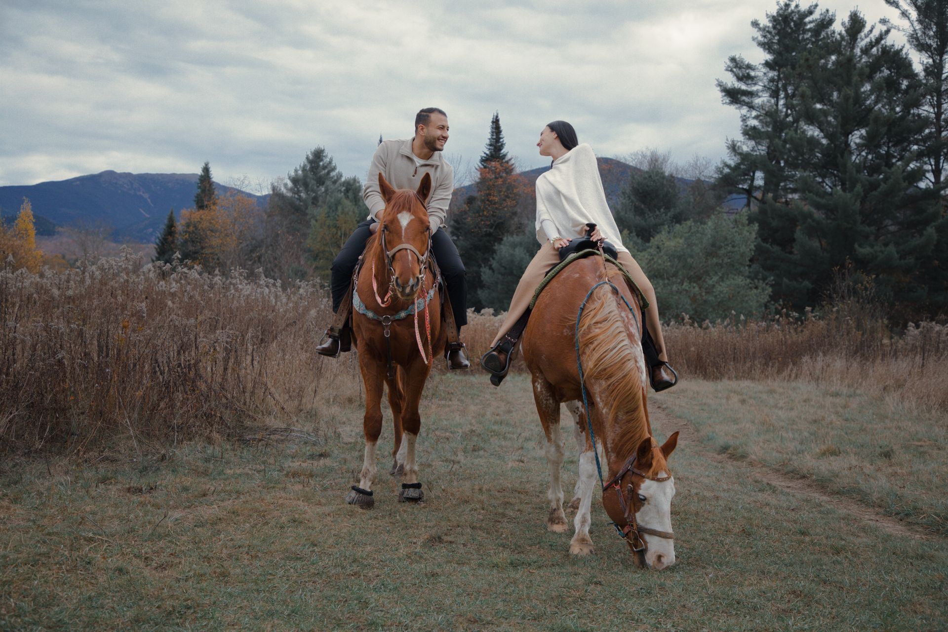 Two people on horseback in a field; one horse grazing. Mountains and trees in the background under an overcast sky.