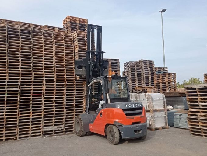 Carrello elevatore arancione in un deposito di legname con pallet e casse di legno impilati.