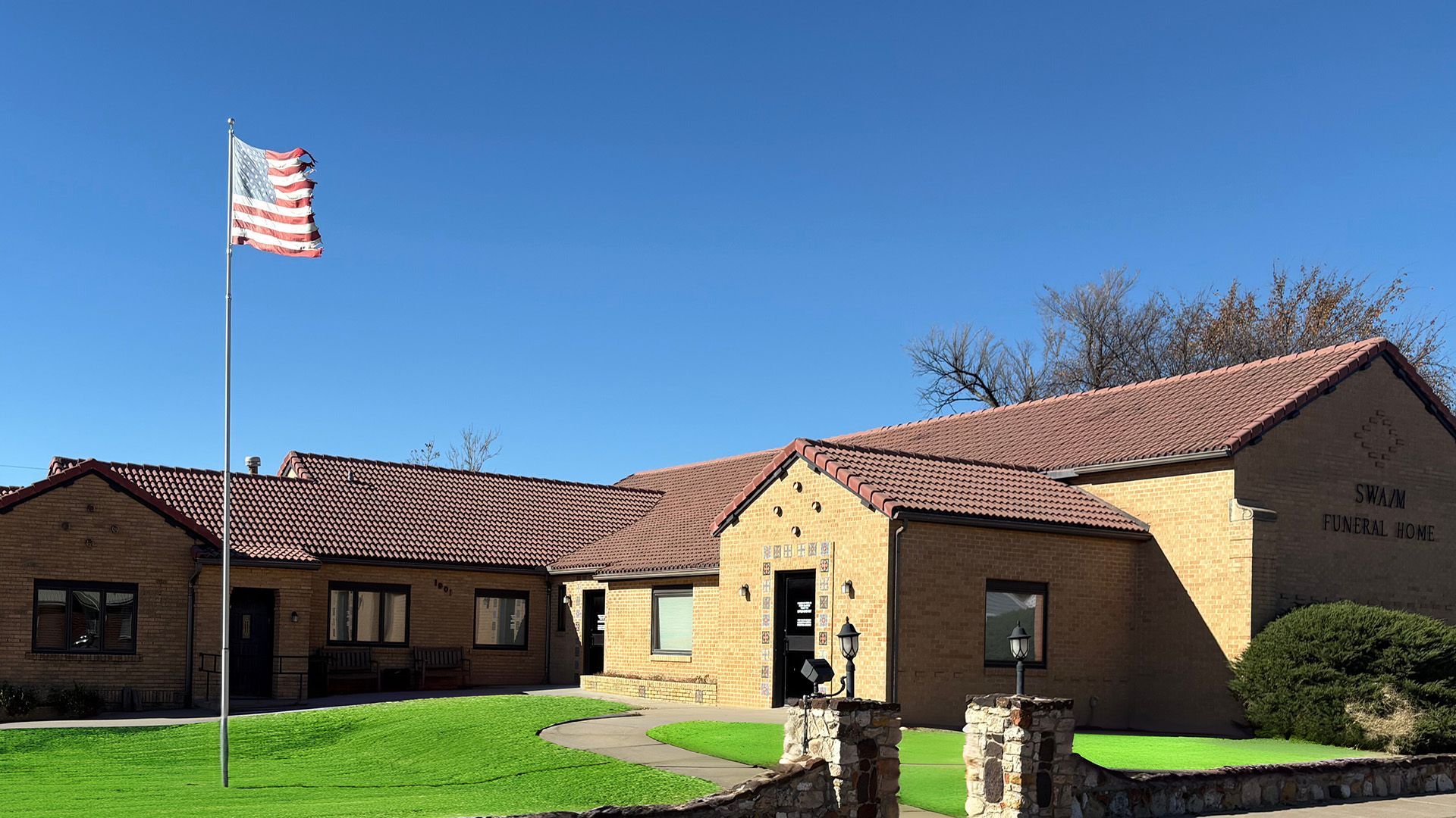 A tan building with a red roof, American flag, and stone wall, in front of green grass and trees.