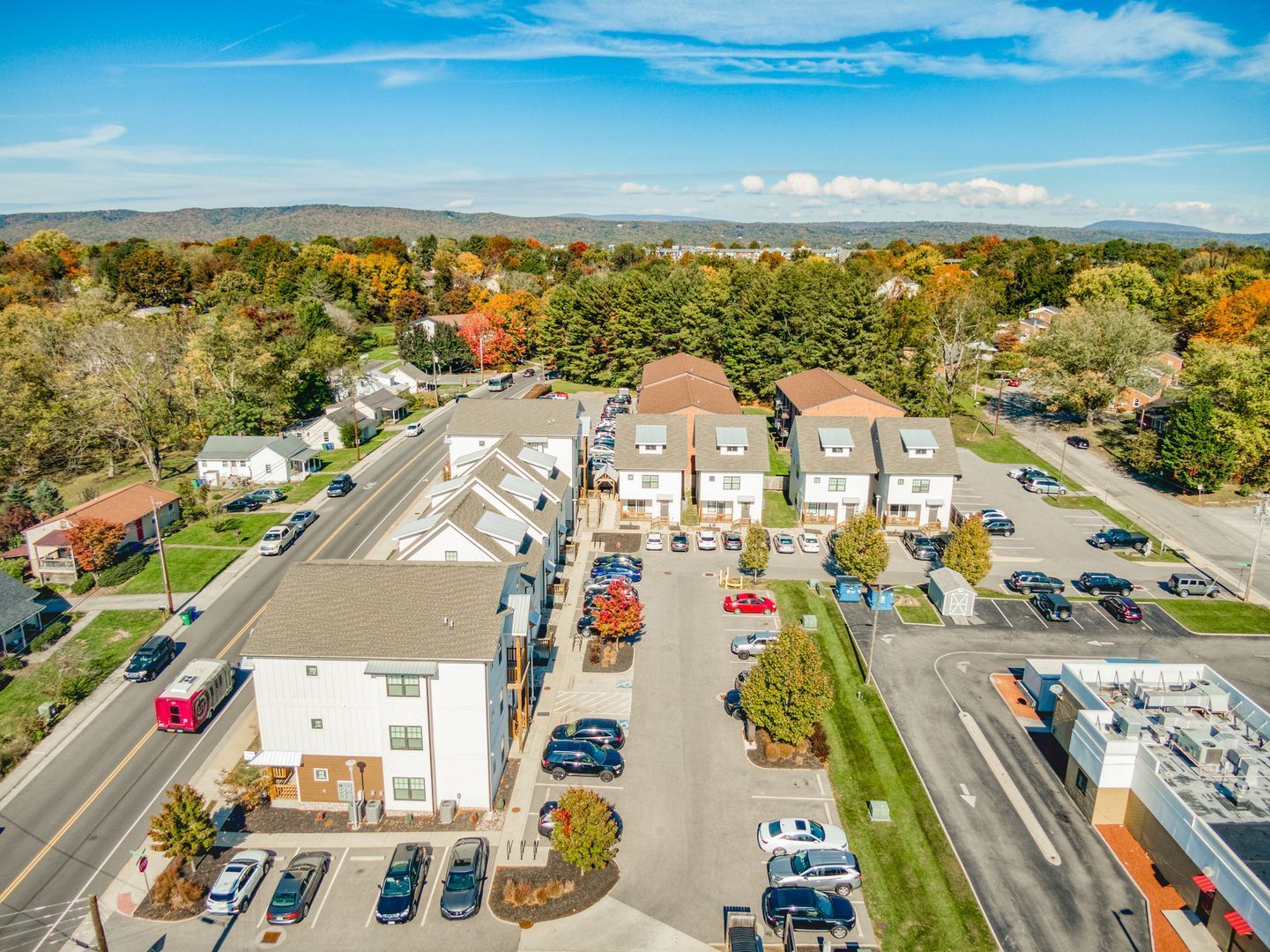 Aerial view of townhouses and parking, lined by road with cars and trees, on a sunny day.