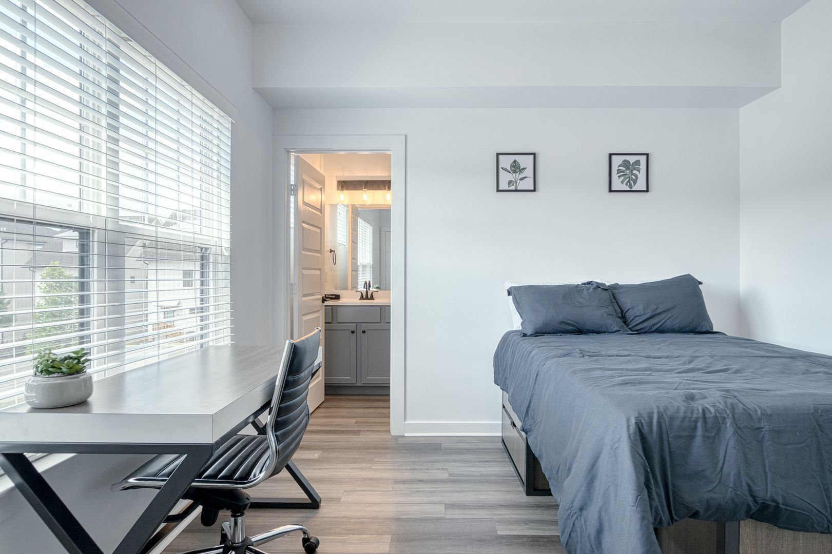 Modern bedroom with a desk, bed, and bathroom access. Light blue bedding and gray desk.