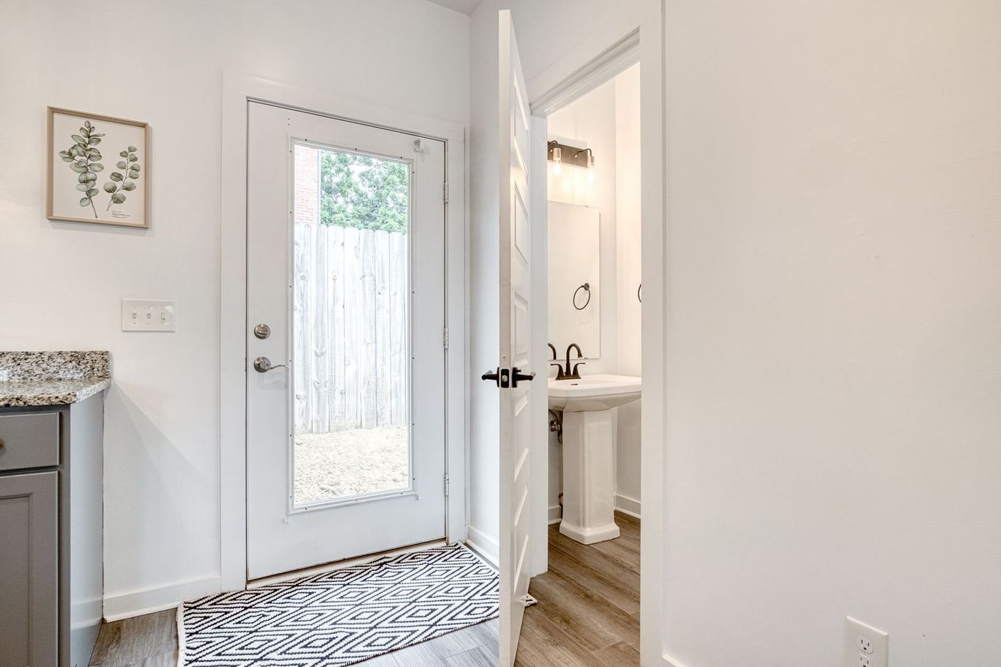 A doorway leads to a backyard and a powder room. White walls, gray countertop, and a patterned rug.