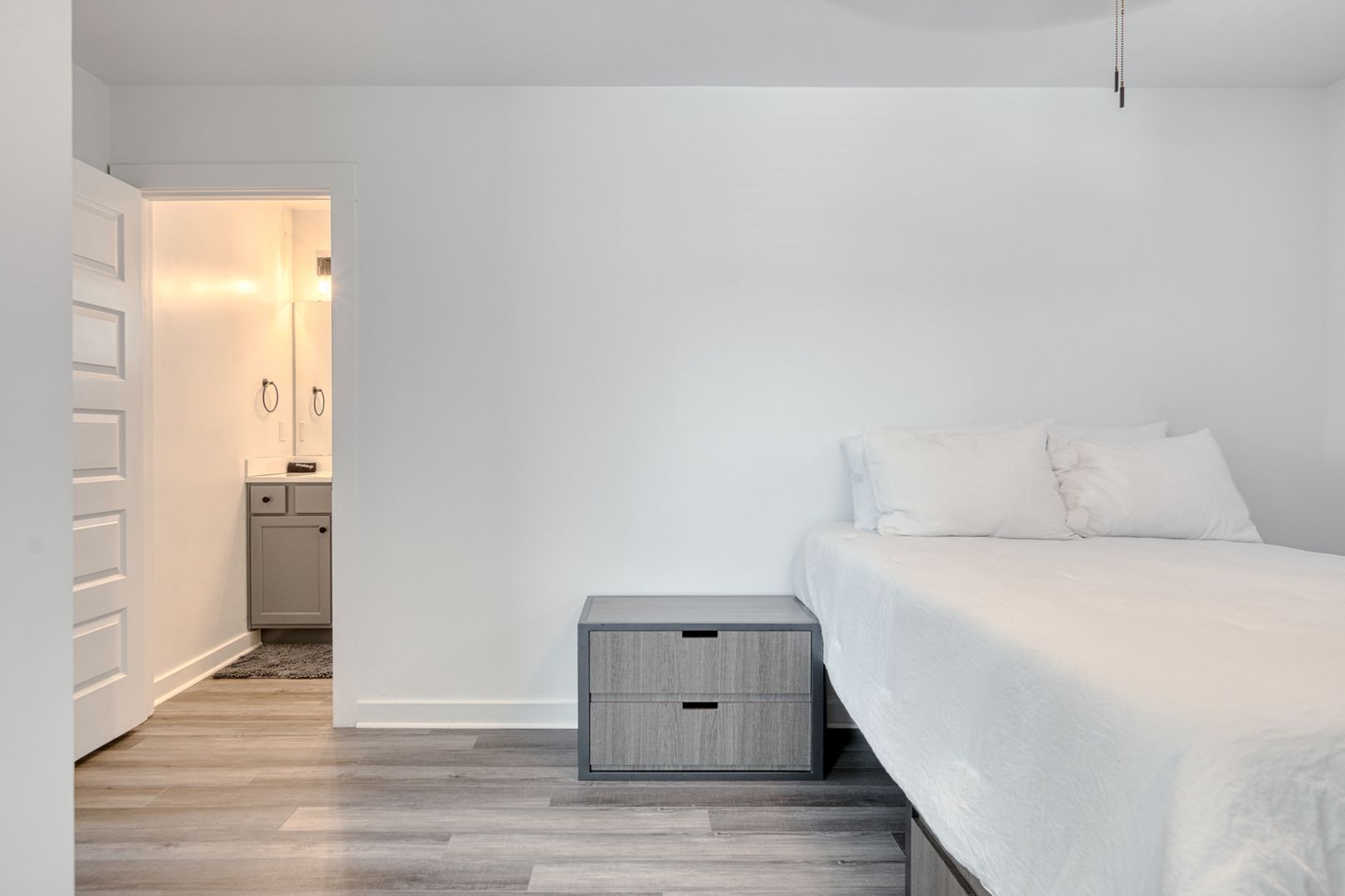Bedroom with bed, nightstand, and open door to a bathroom. White walls and gray wood flooring.
