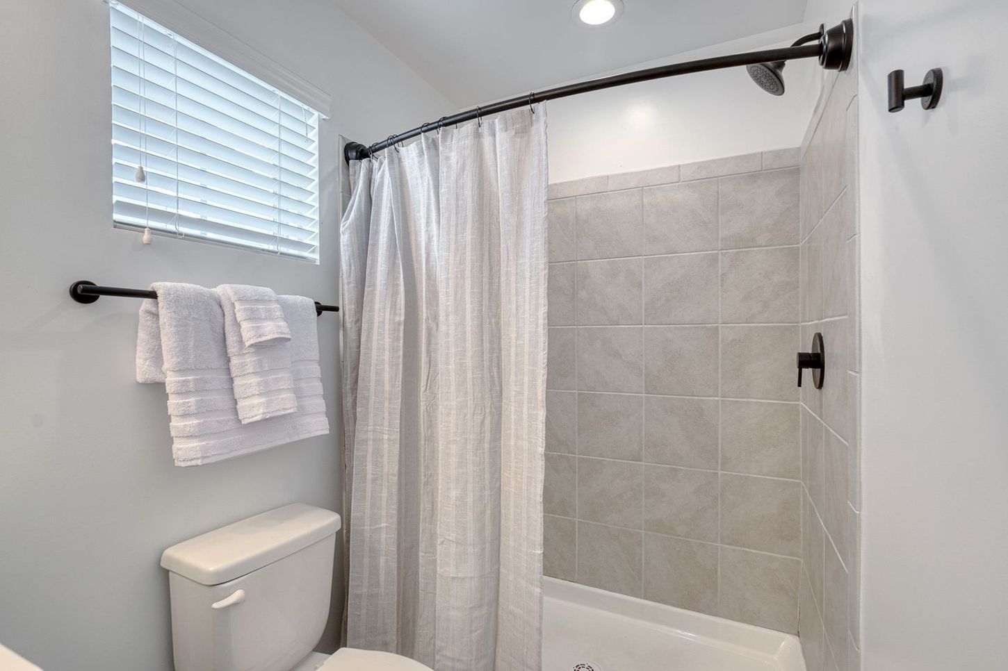 Bathroom with a toilet, shower, towels, and a window with blinds. Gray and white color scheme.