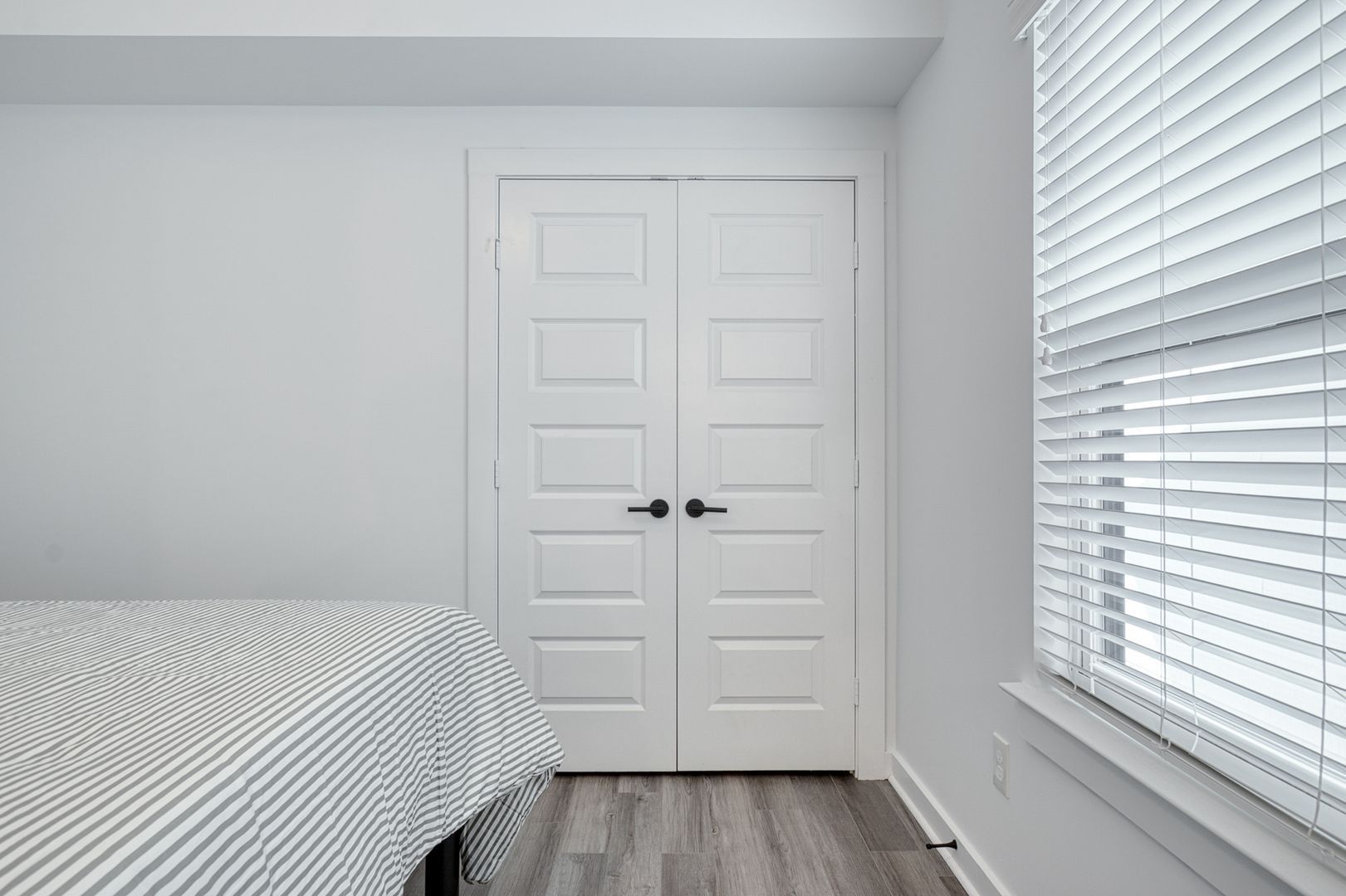 Bedroom with white doors, blinds, and striped bedsheet. Grey floor, white walls.