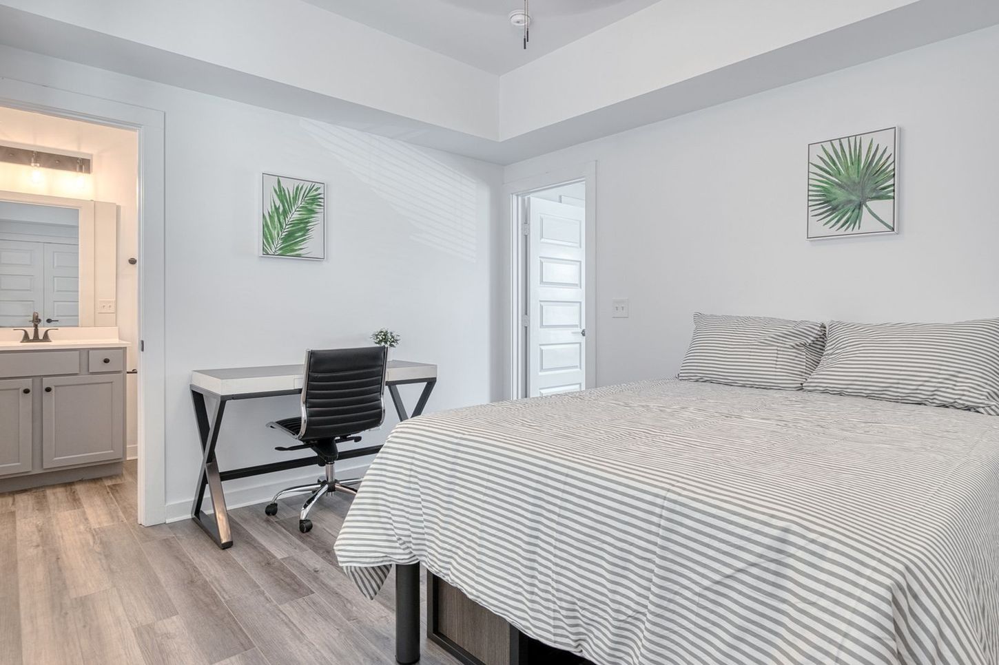 Bedroom with a bed, desk, and a partially visible bathroom. White walls, wood-look floor, and two green leaf art prints.