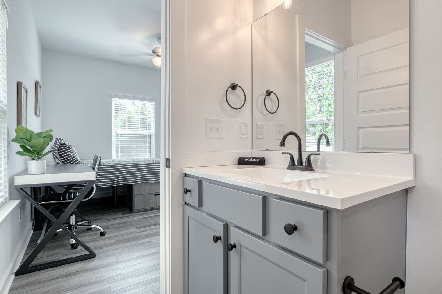 Bathroom with gray vanity, black fixtures, and a view into a bedroom with a desk.