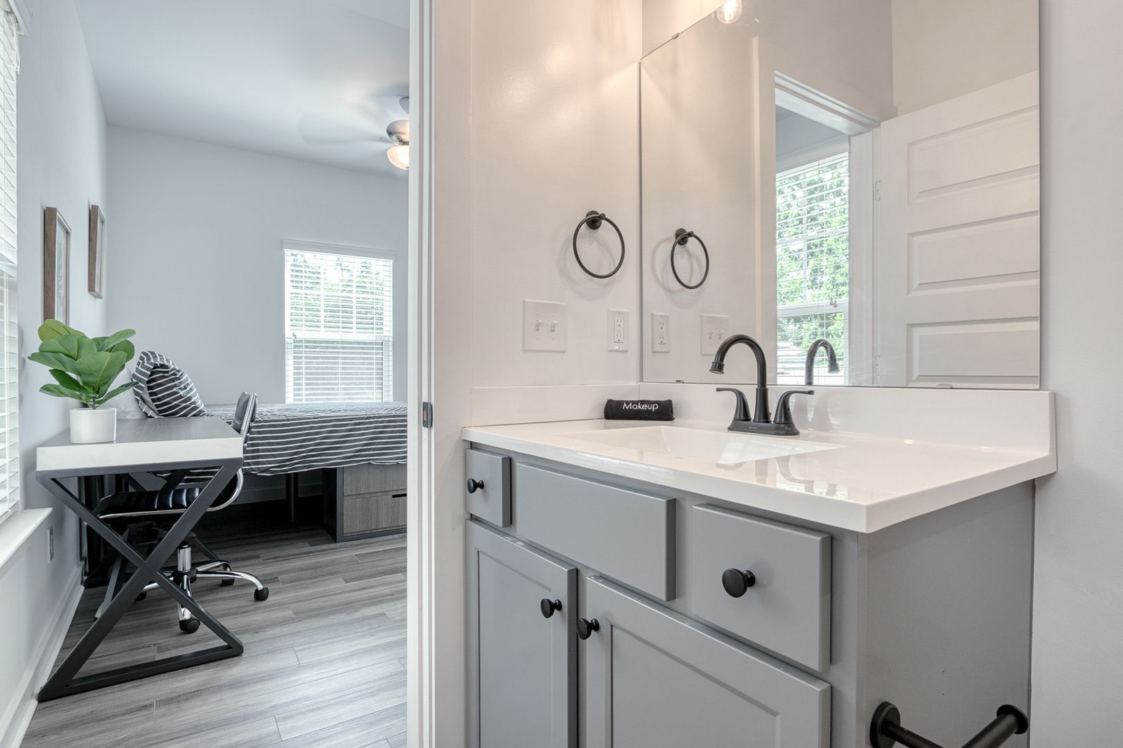 Bathroom with gray vanity, black fixtures, and a view into a bedroom with a desk.