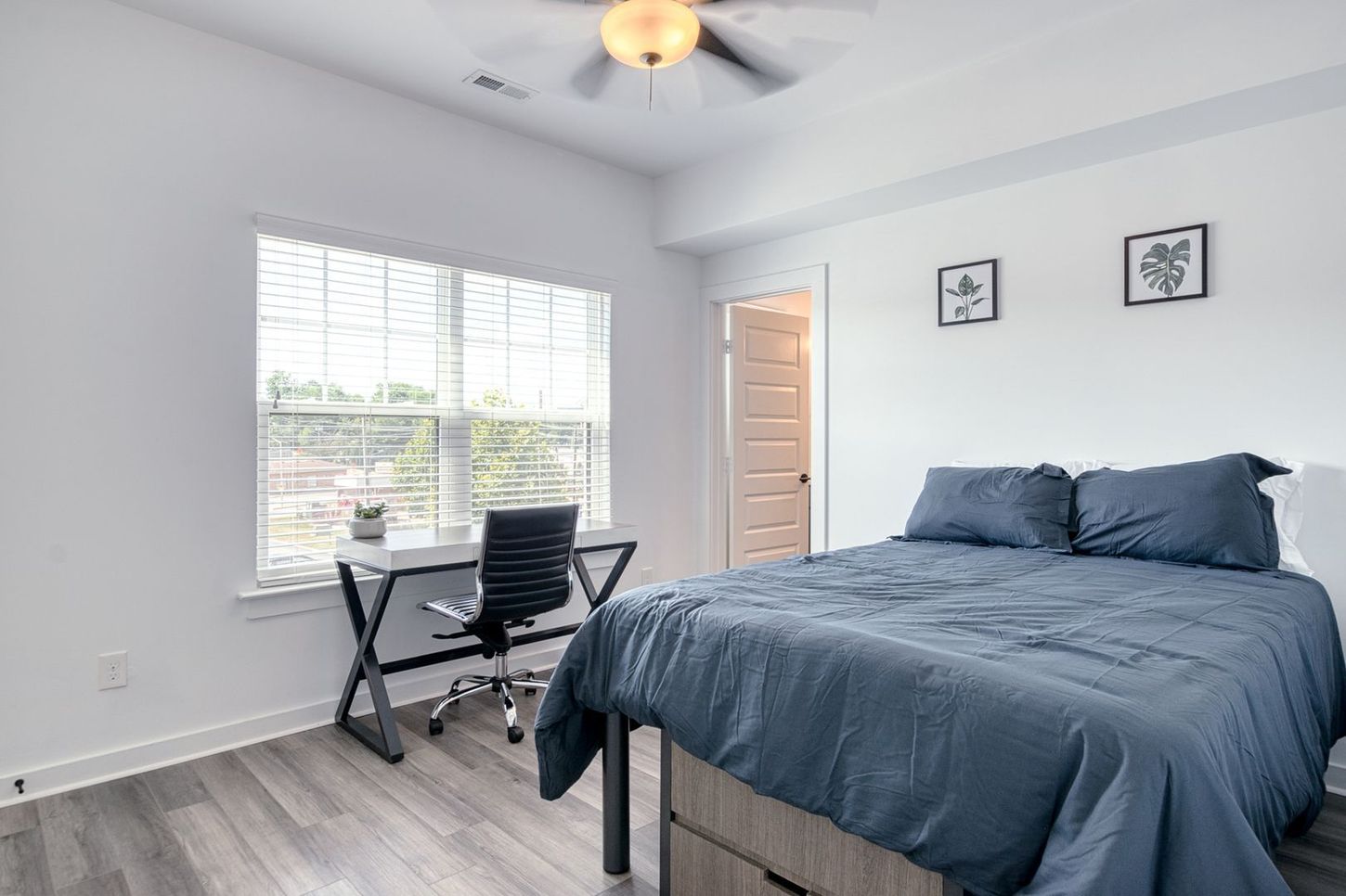 Bedroom with blue bedding, desk, window, and two framed prints on a white wall.