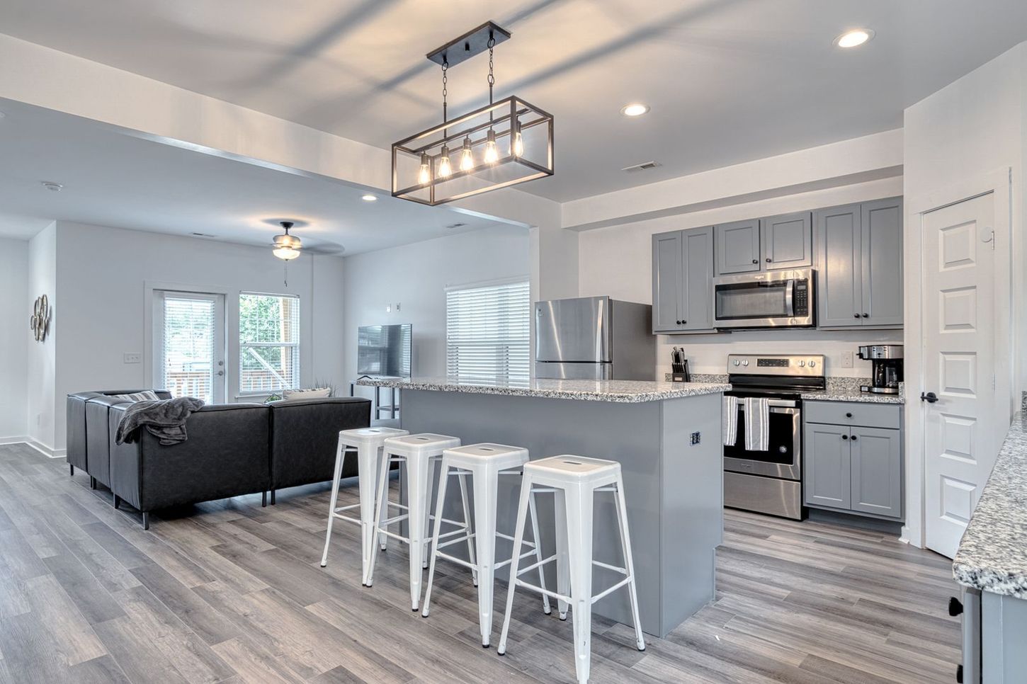 Modern open-concept kitchen and living room with gray cabinets, stainless steel appliances, and white bar stools.