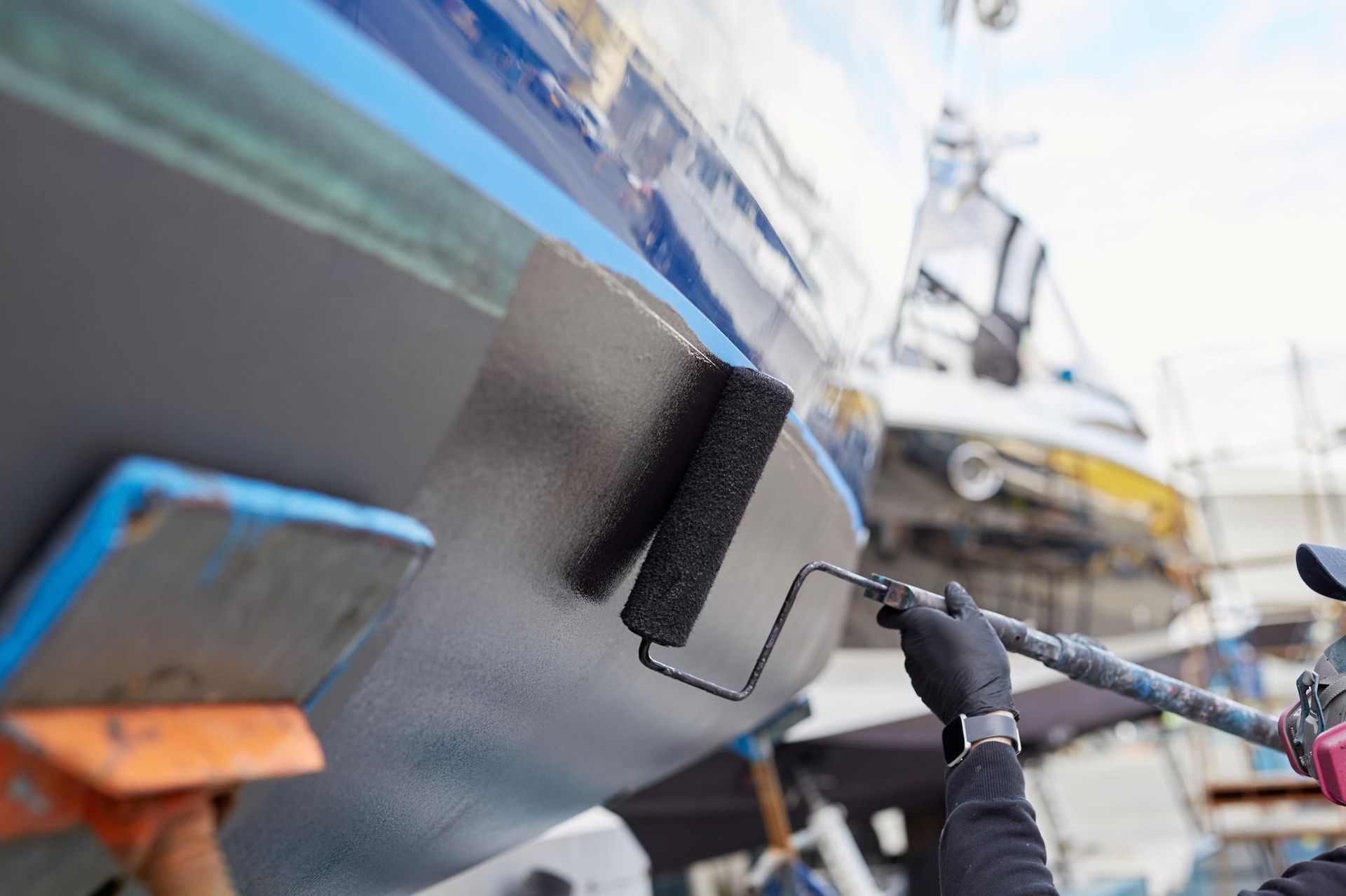 Person paints the hull of a boat with a roller; blue, black, and white colors are visible.