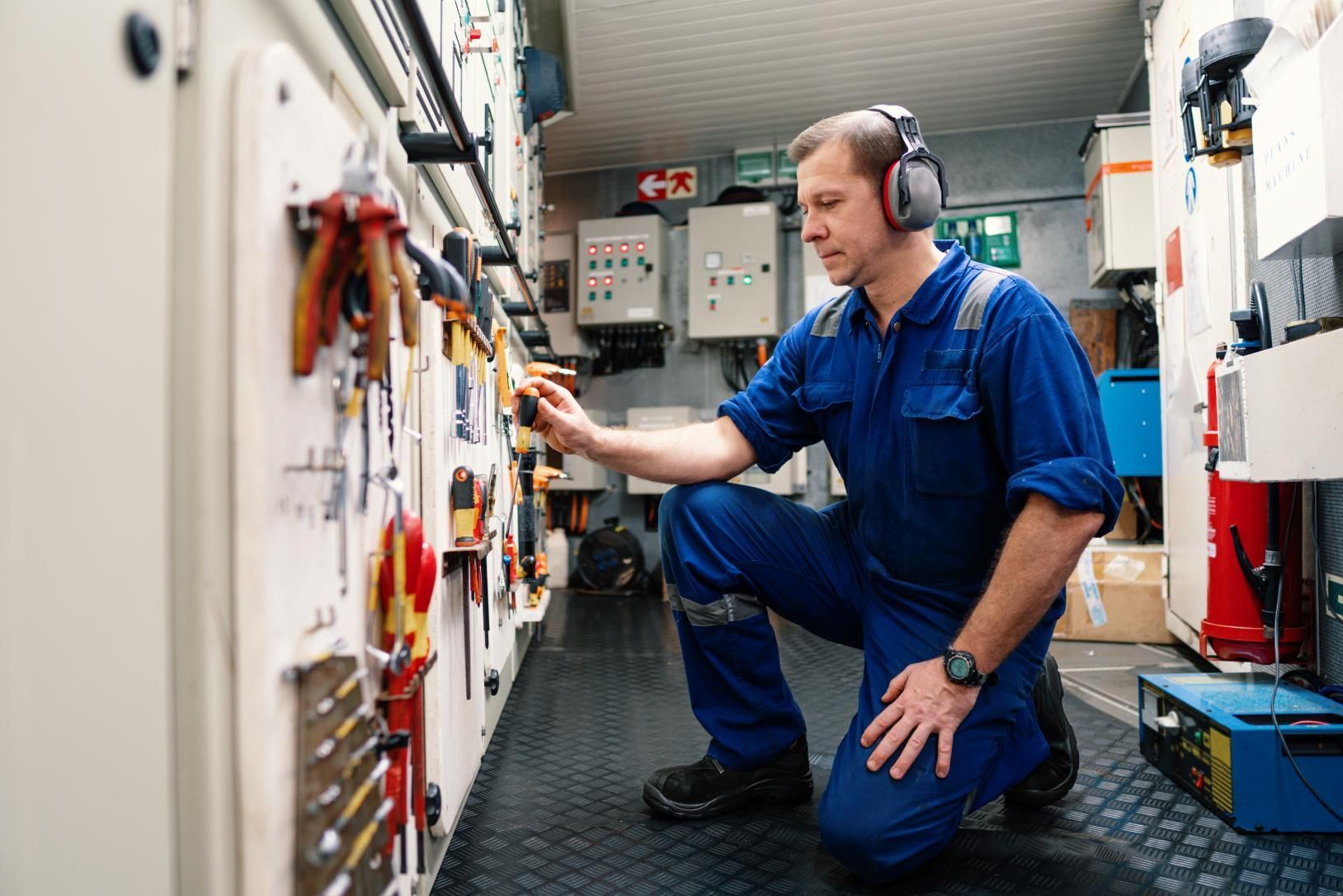 Mechanic kneeling, wearing ear protection, in an equipment room, inspecting tools on a wall.